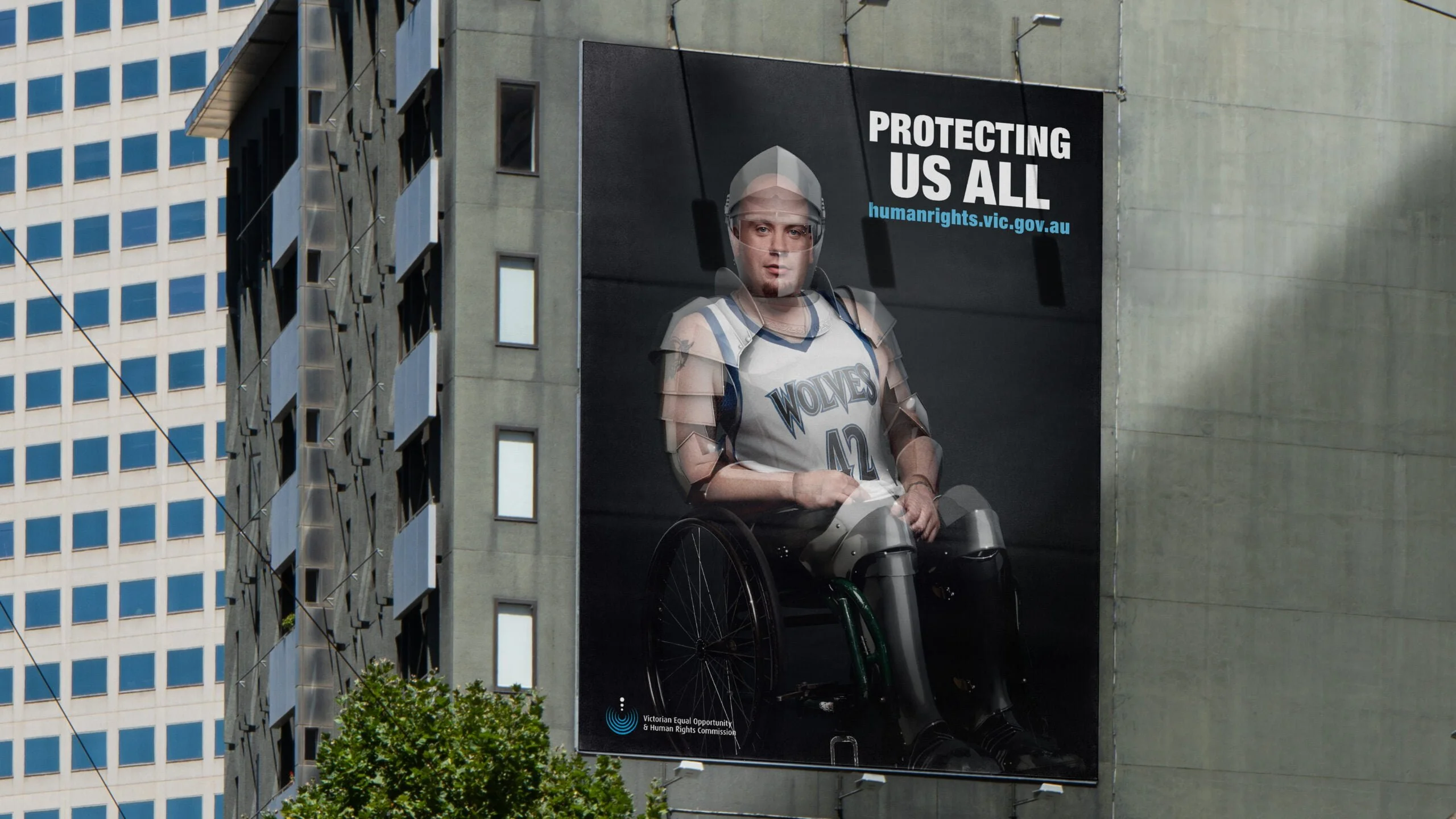 Building-side billboard: 'Protecting us all', a man in a Wolves basketball singlet in a wheelchair wearing translucent armour