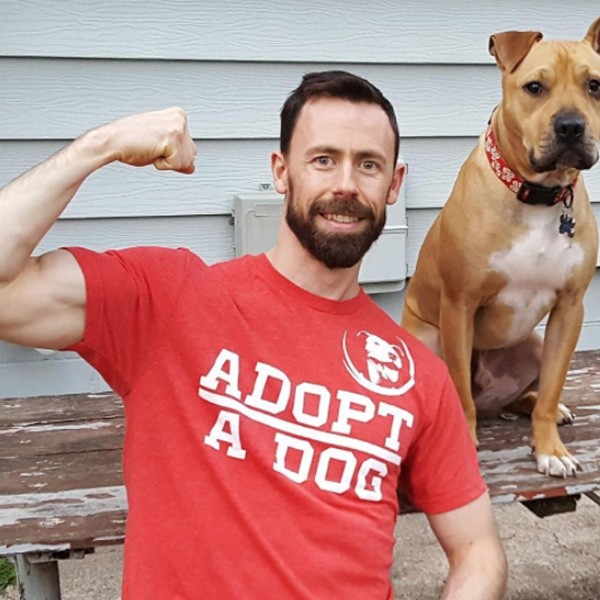 Man flexing bicep in "Adopt a Dog" shirt with dog sitting nearby.