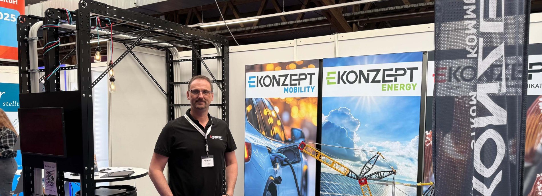 Mann posiert auf Messe vor "Ekonzept"-Stand mit Mobilitäts- und Energie-Werbung, Hintergrund mit Ausstellungsstruktur