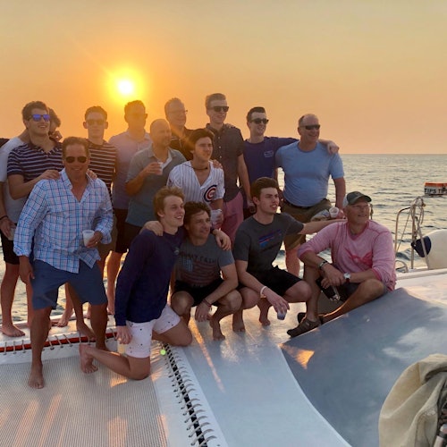 A group of 14 people pose for a photo on a boat at sunset, with a calm sea in the background.