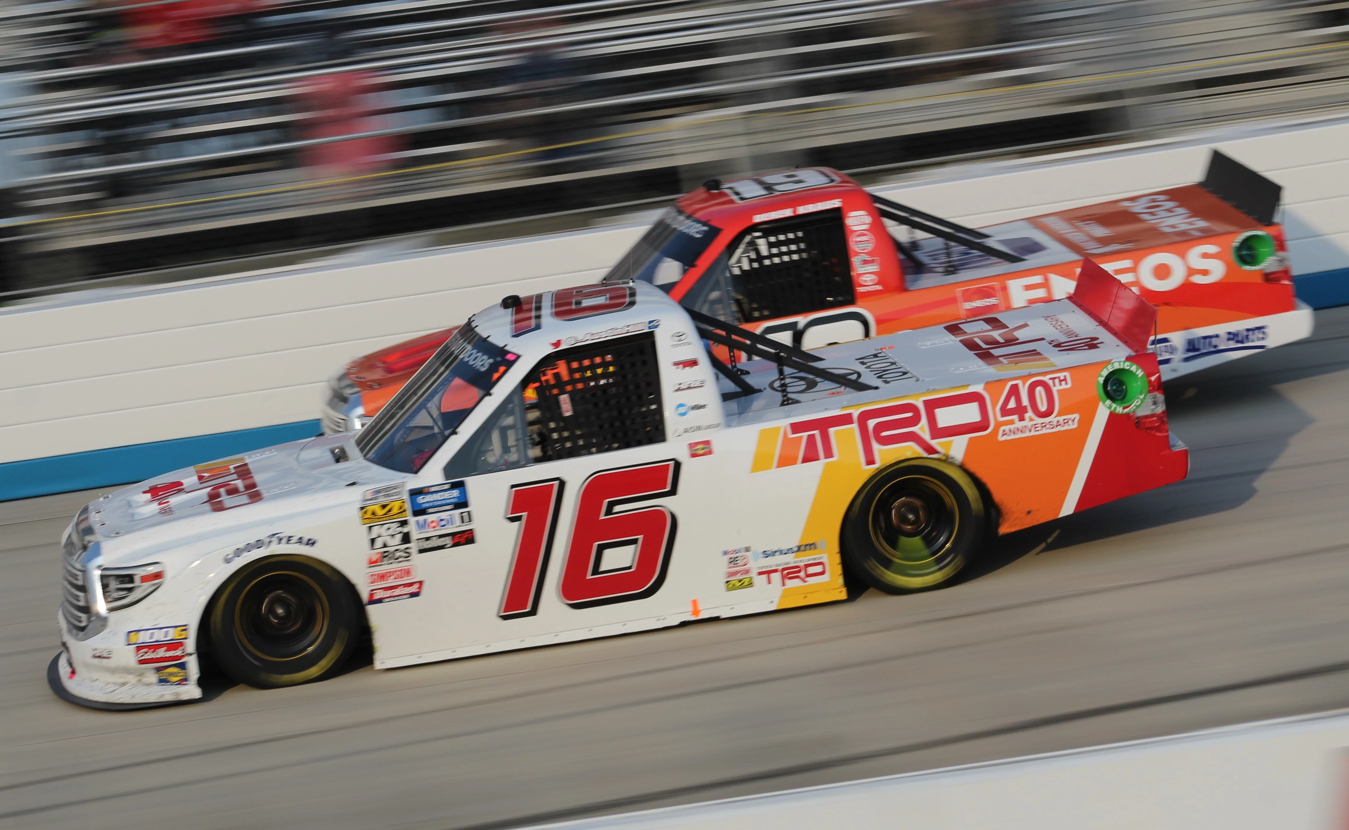Two race trucks on track during a NASCAR event, with the front vehicle featuring a large TRD 40th Anniversary logo in red and yellow on a white Toyota body. Highlights TRD’s legacy and involvement in professional motorsports.