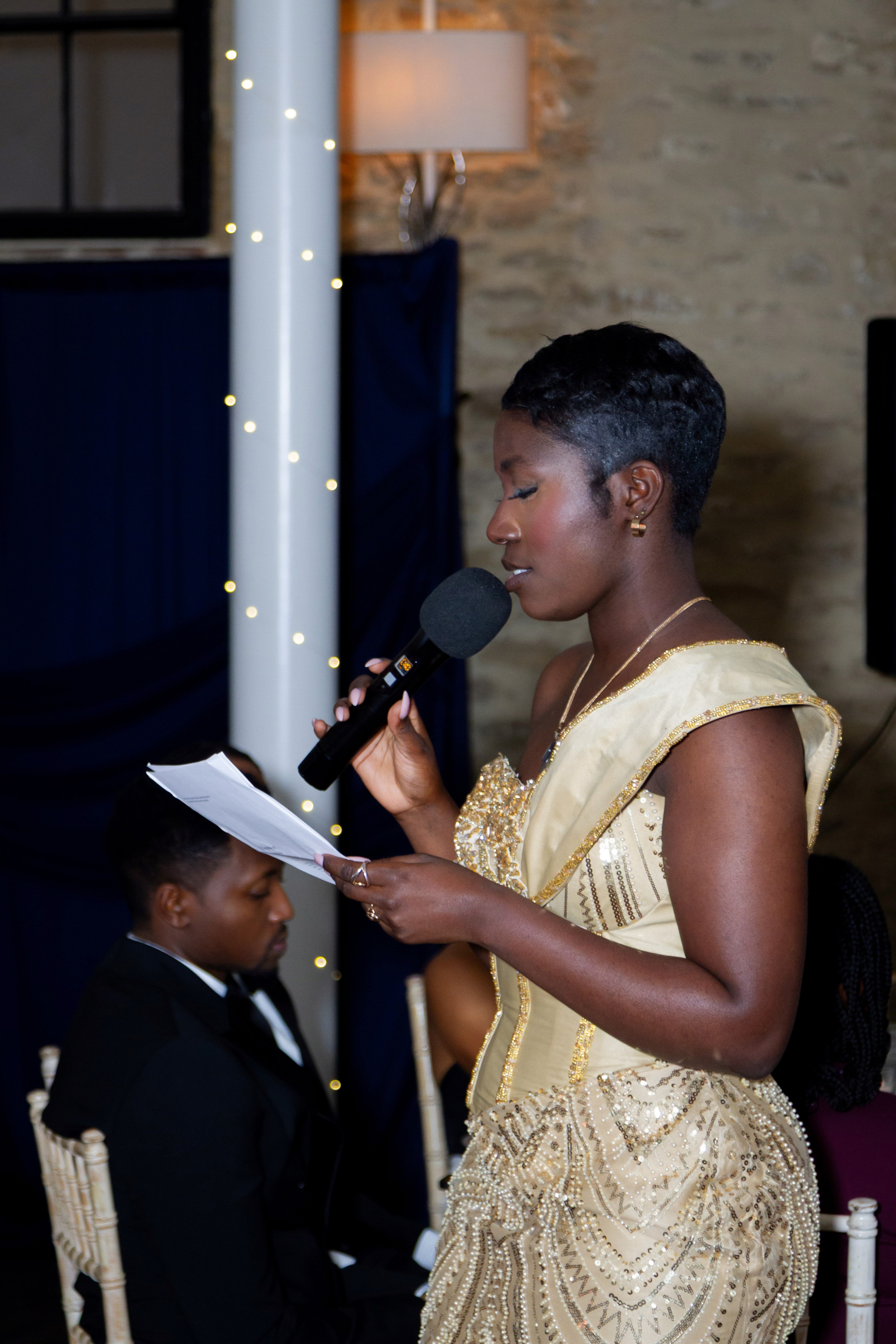 A woman in a formal dress speaks into a microphone, reading from a paper.