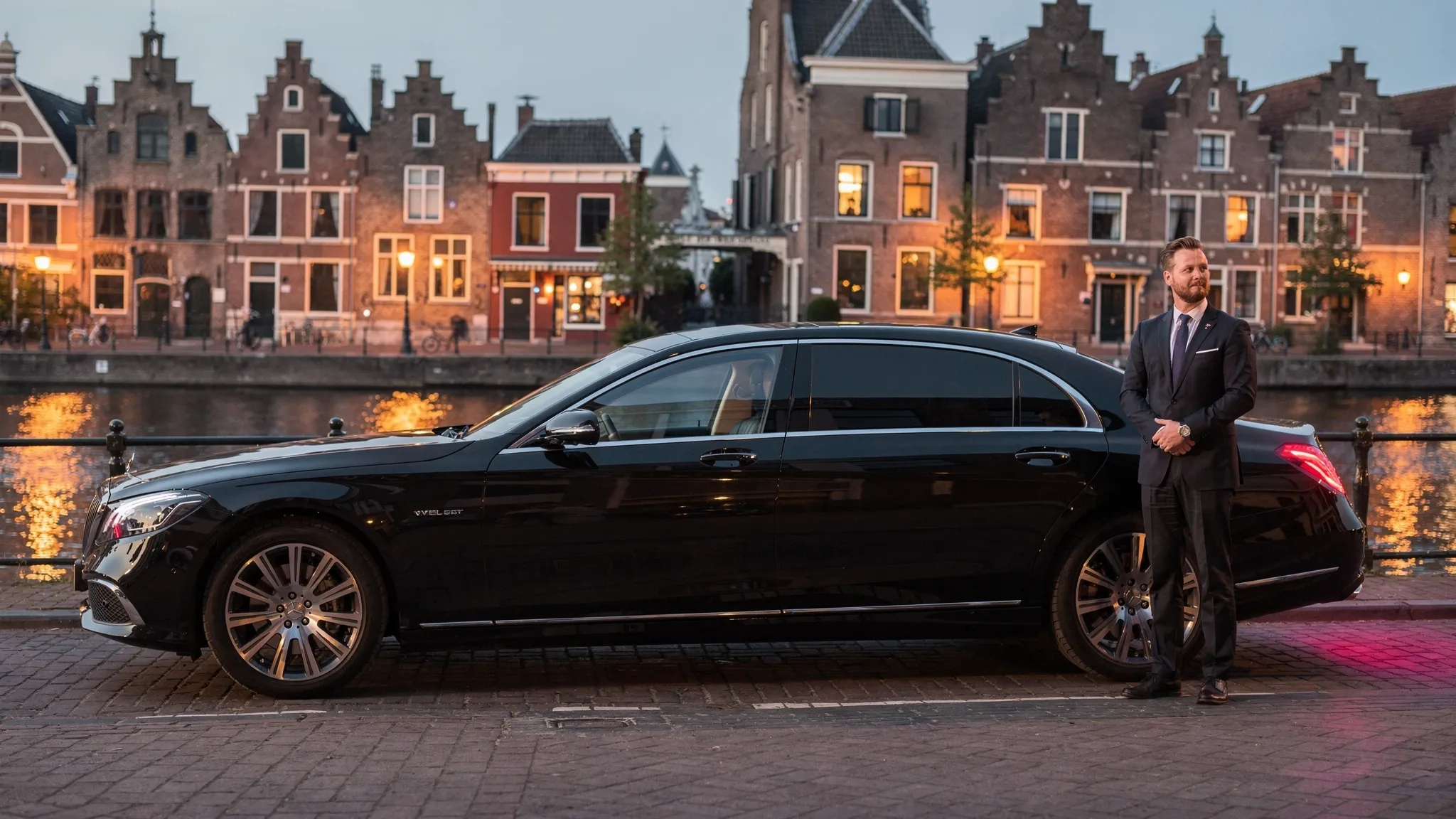 Een representatieve zwarte chauffeursauto staat klaar bij een stijlvolle entree in Alkmaar, met op de achtergrond historische gevels en avondlicht. De chauffeur wacht discreet naast de auto terwijl de omgeving rustig en professioneel oogt.