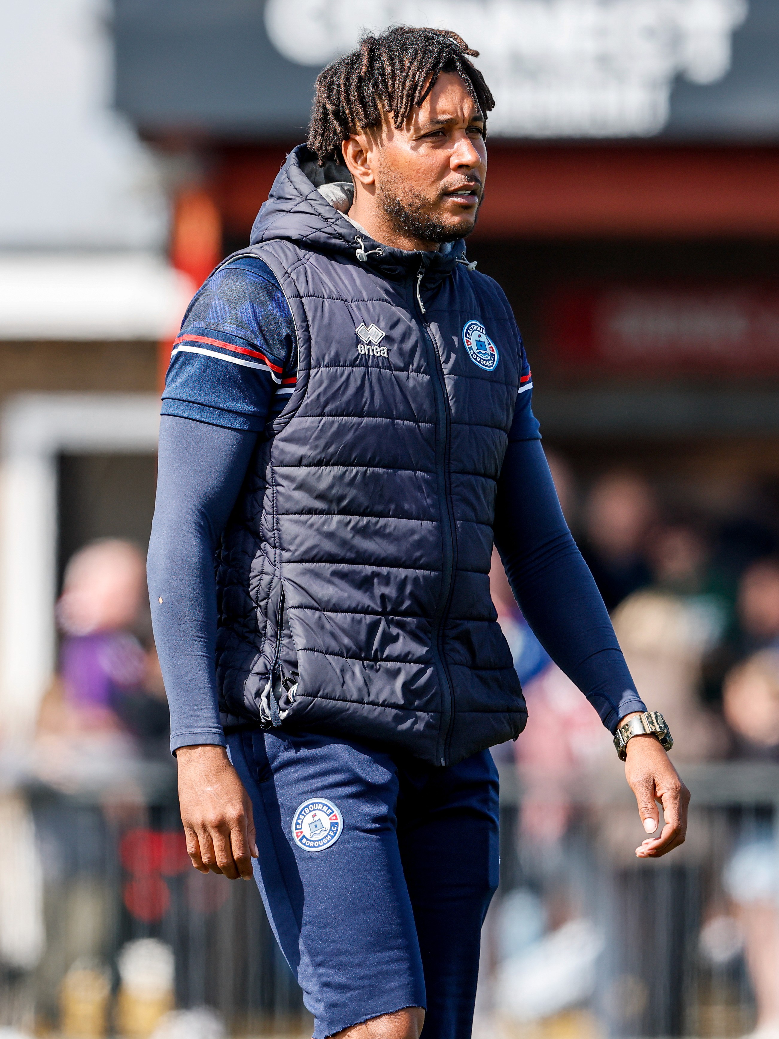 A coach in a padded vest and athletic gear stands on the sidelines during a bright day, focusing on the game, with a blurred background of a stadium and spectators.