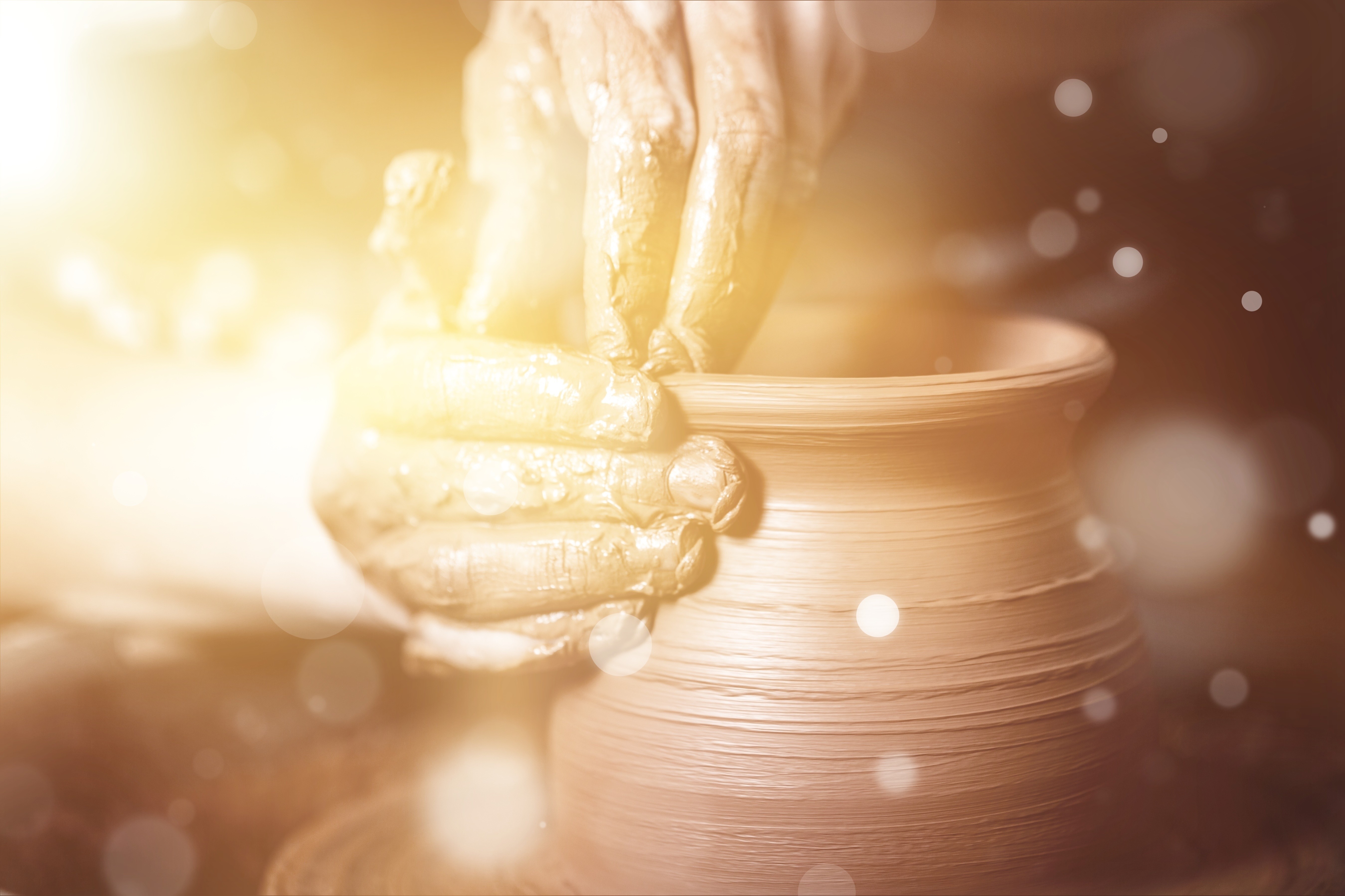 Close-up of hands actively shaping a wet clay pot spinning on an artisan's pottery wheel, surrounded by warm lighting and splashing water droplets.