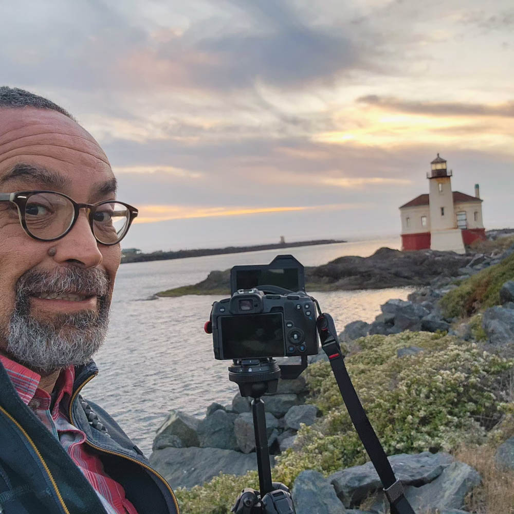 Benjamin Hernandez standing by a lighthouse with his camera on a tripod at sunset.