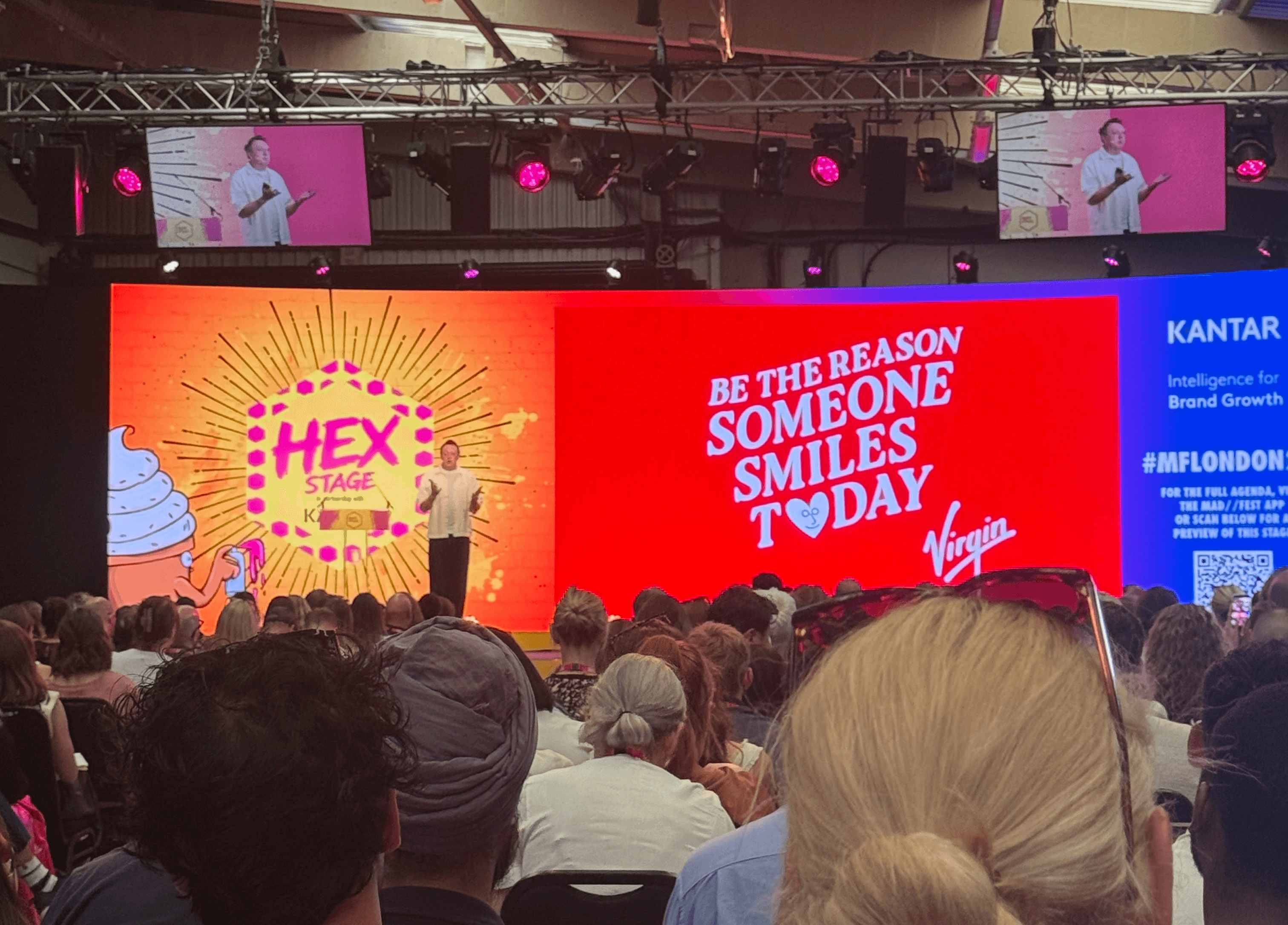 A speaker presents on the Hex Stage in partnership with Kantar, with a red screen reading 'Be the Reason Someone Smiles Today' and the Virgin logo, viewed over a crowded audience.