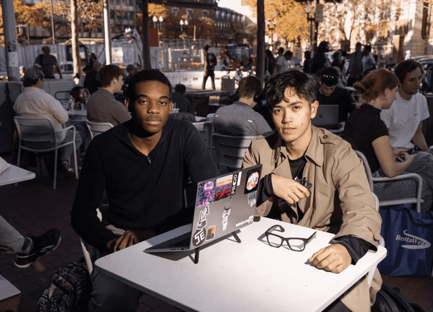 Two students looking at a camera sitting at a table, Meta Ray Bans AI Glasses in view on the table.