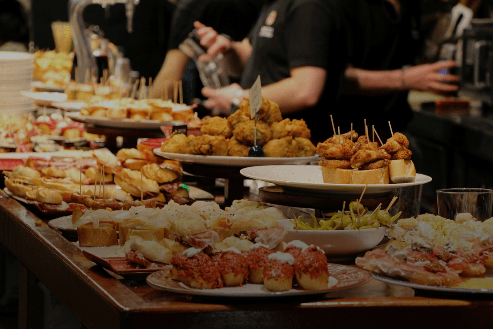 A long bar counter displays various pintxos (Basque tapas). The pintxos include seafood, meat, and vegetables.