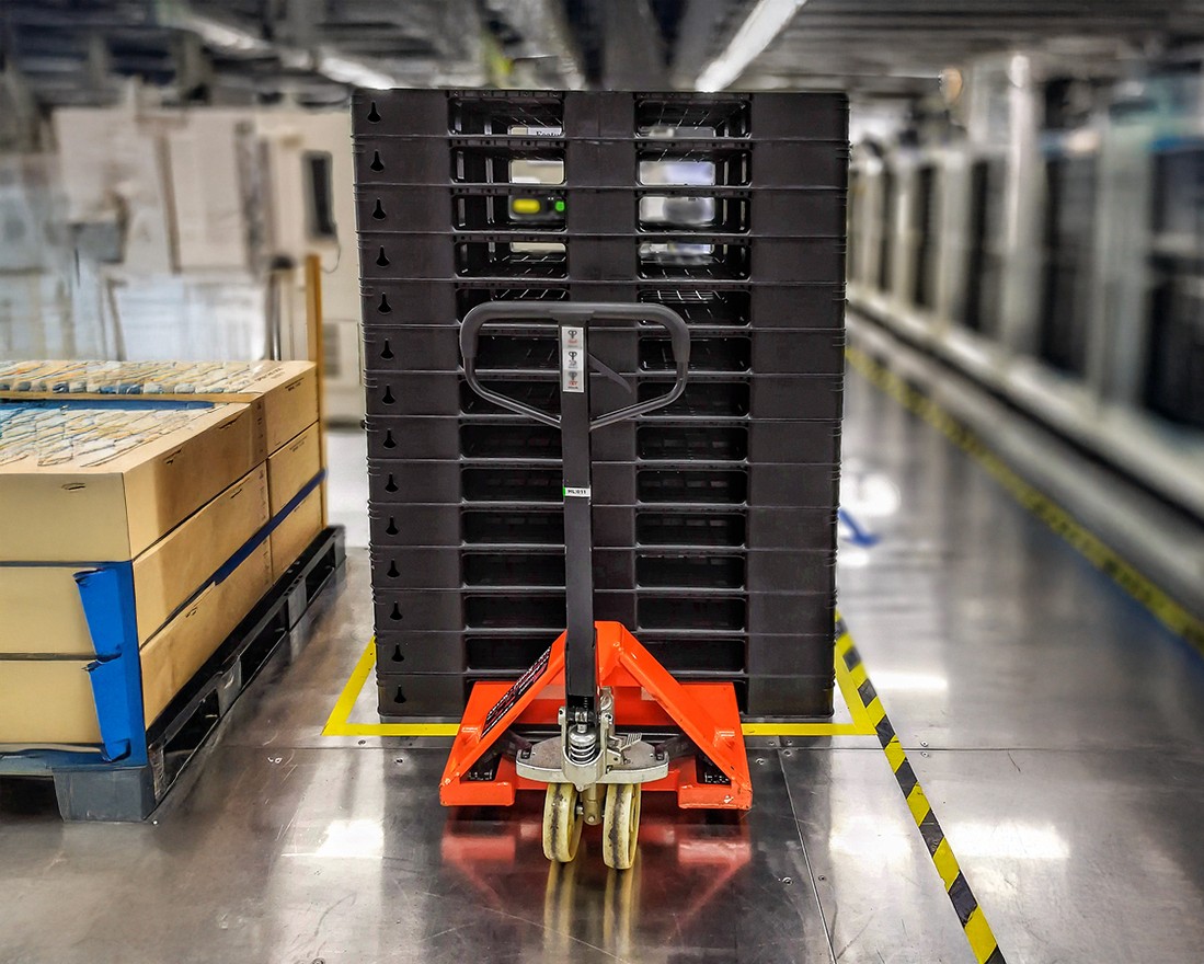 Orange forklift holding many black platic pallets in an industrial warehouse setting