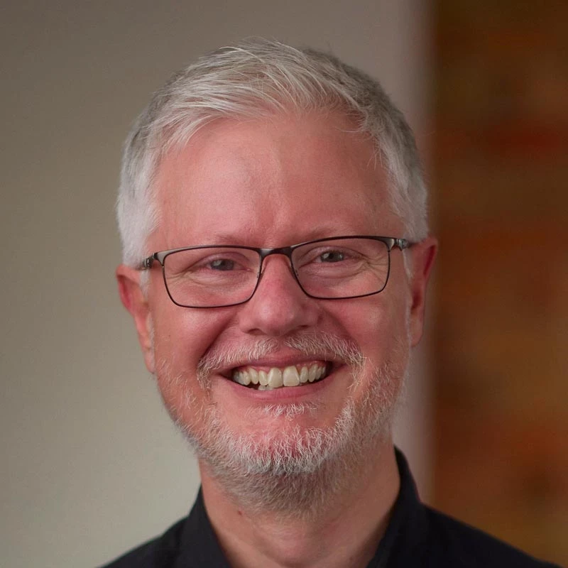 A person with short grey hair wears a dark shirt in a softly lit indoor setting. A warm-toned background wall is partially visible.