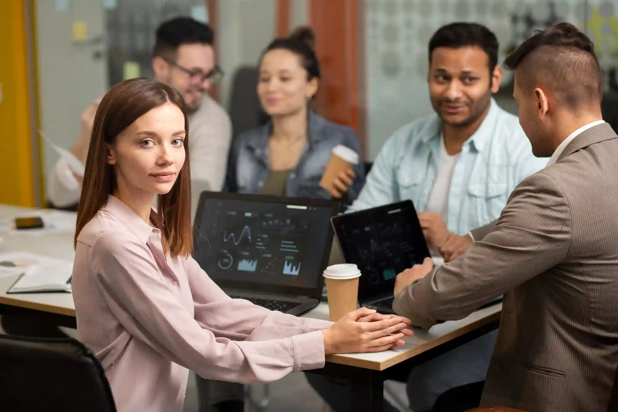 A diverse team of professionals engaging in a collaborative business meeting with laptops and data analytics on screen.