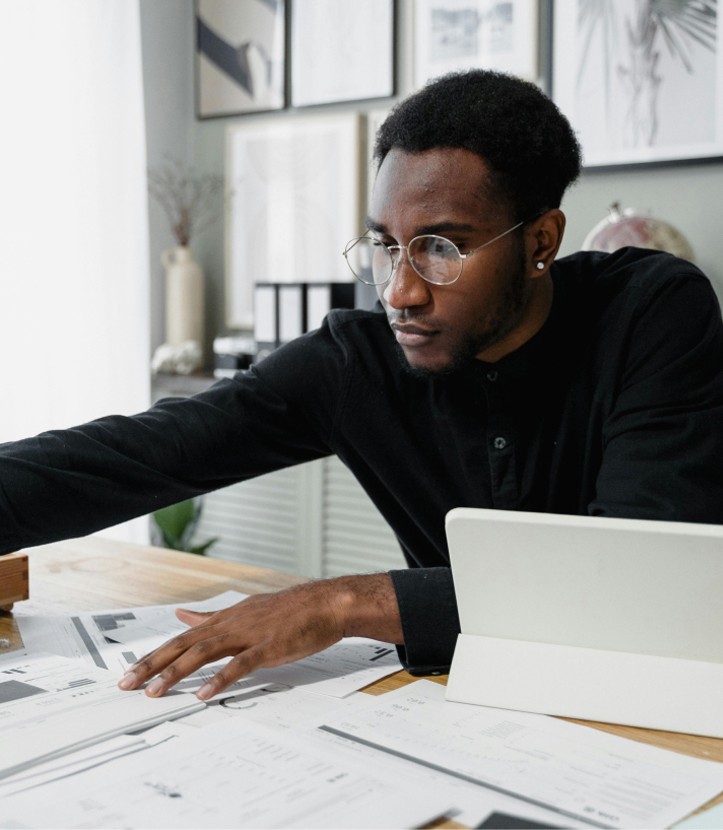 Focused professional man wearing glasses and a black shirt, working on design or architectural plans at a desk with a tablet and documents.