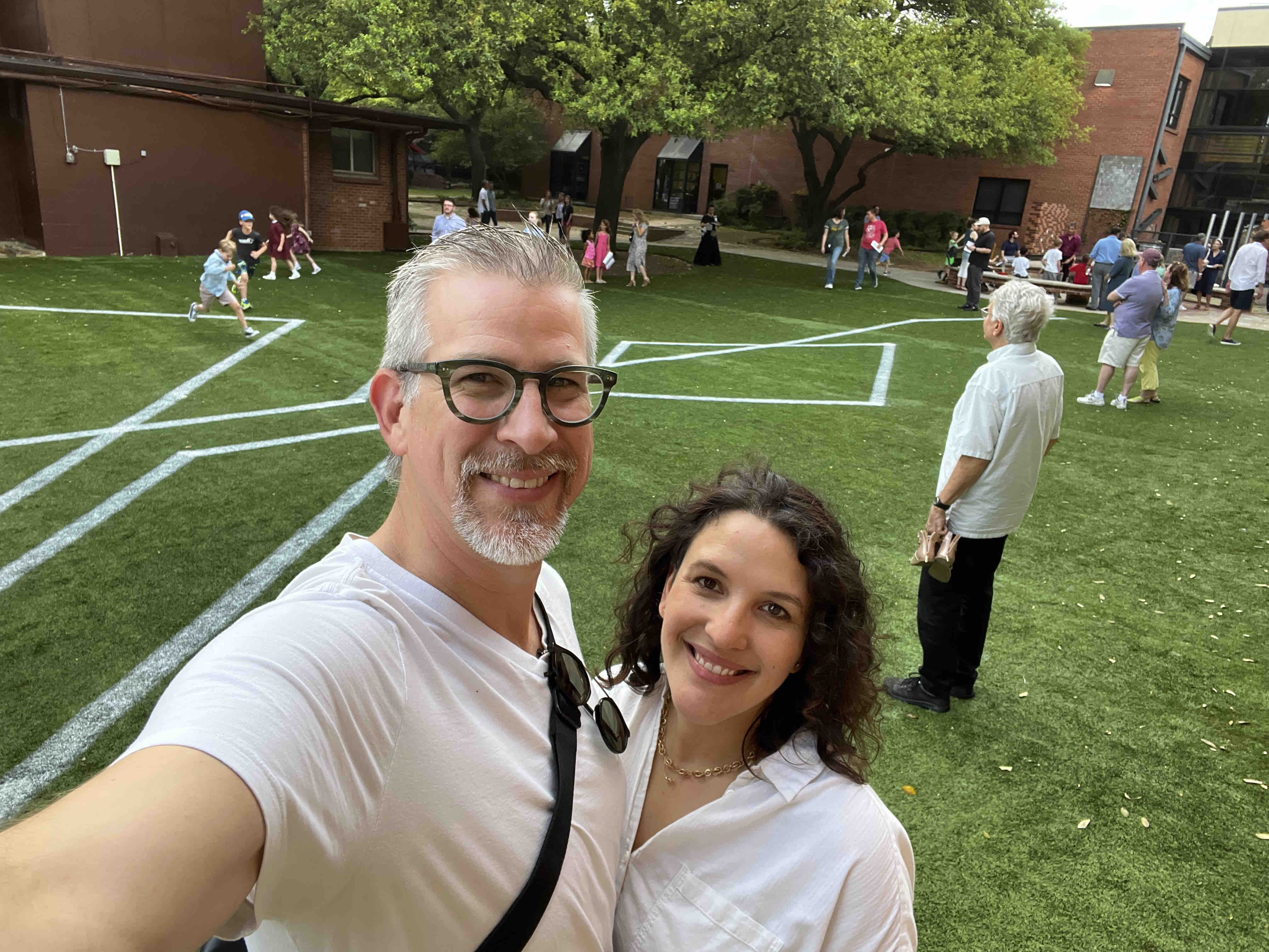A smiling man and woman take a selfie outdoors, with people playing sports in the background.