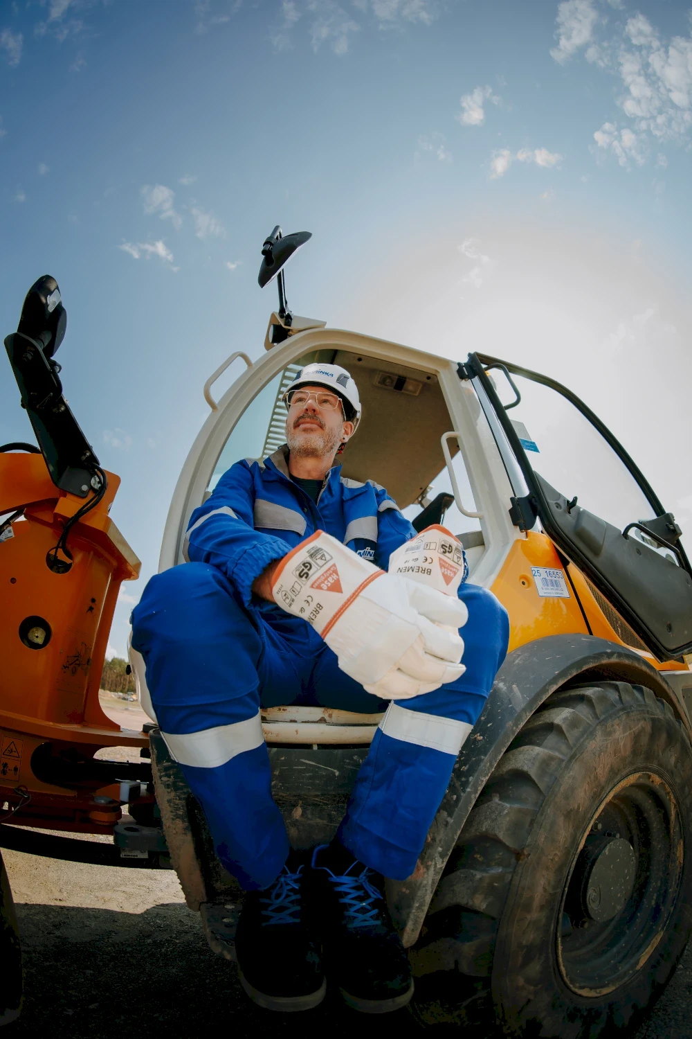 Ein Arbeiter sitzt auf einem Baufahrzeug. Er trägt einen blauen Overall, Handschuhe und einen Helm. Blauer Himmel.