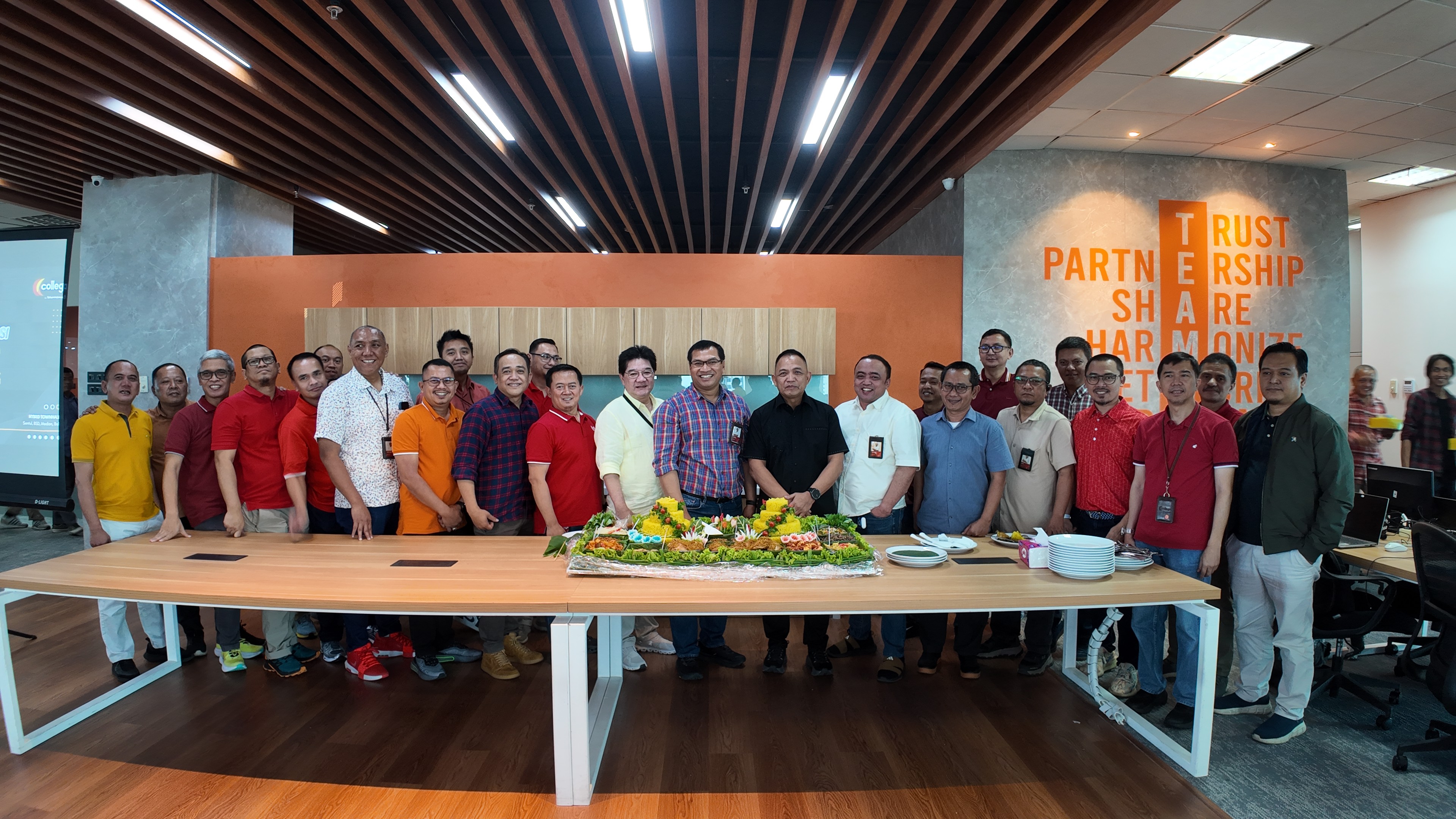 Foto bersama sekelompok karyawan di ruang kantor modern setelah acara syukuran, berdiri berjajar di belakang meja panjang dengan tumpeng nasi kuning di tengah, sambil tersenyum dan menunjukkan gestur kebersamaan di bawah dekorasi dinding bertuliskan nilai perusahaan.