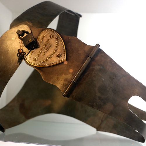 חגורת צניעות ממתכת עם מנעול בצורת לב שעליו חרוט טקסט, מוצגת על מעמד תצוגה.