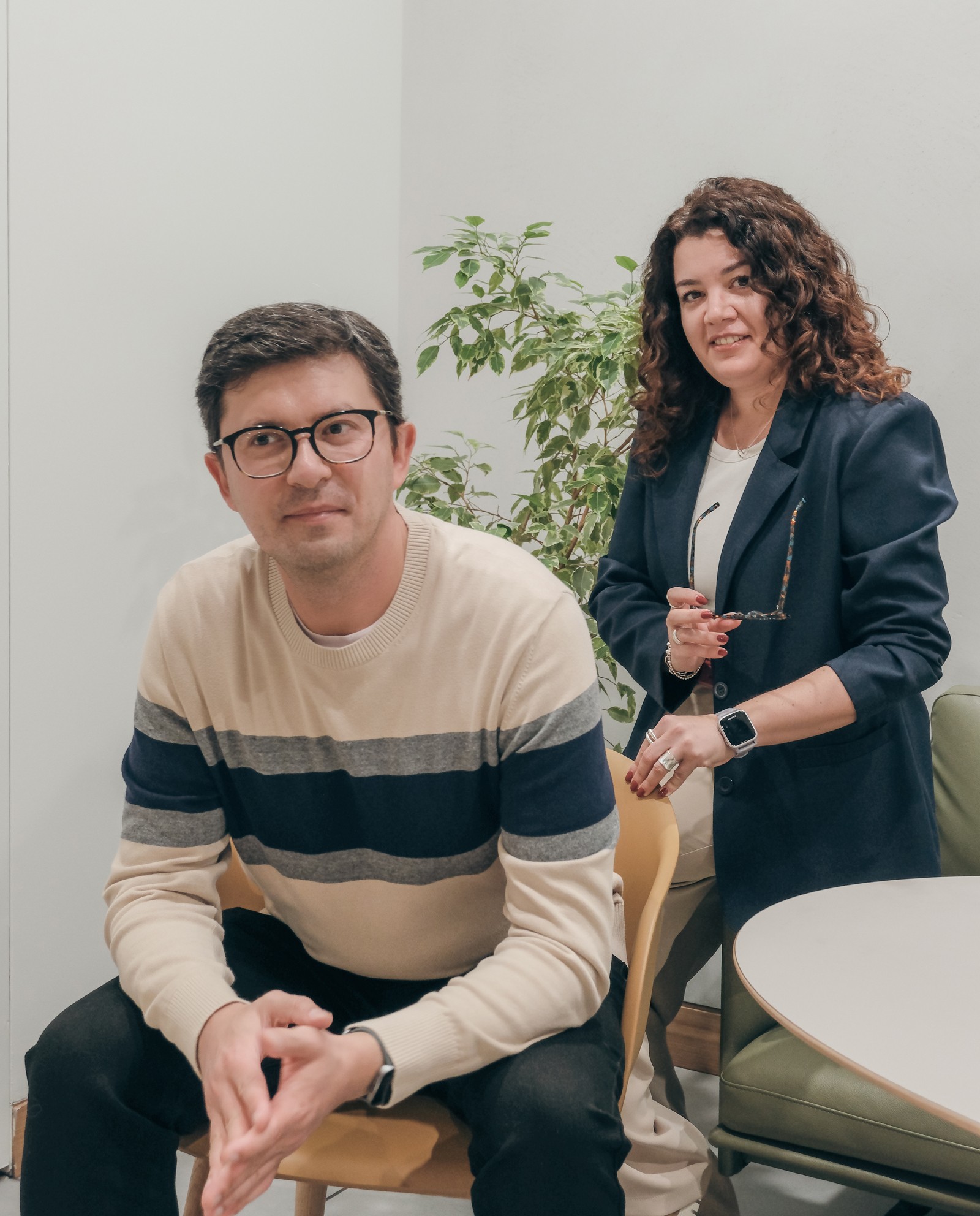 Two colleagues posing in a modern office setting, smiling and looking at the camera.