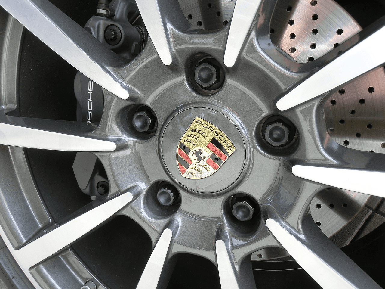 Nahaufnahme einer Porsche Alufelge mit Radschrauben und Porsche Wappen im Zentrum, im Hintergrund die Bremsscheibe