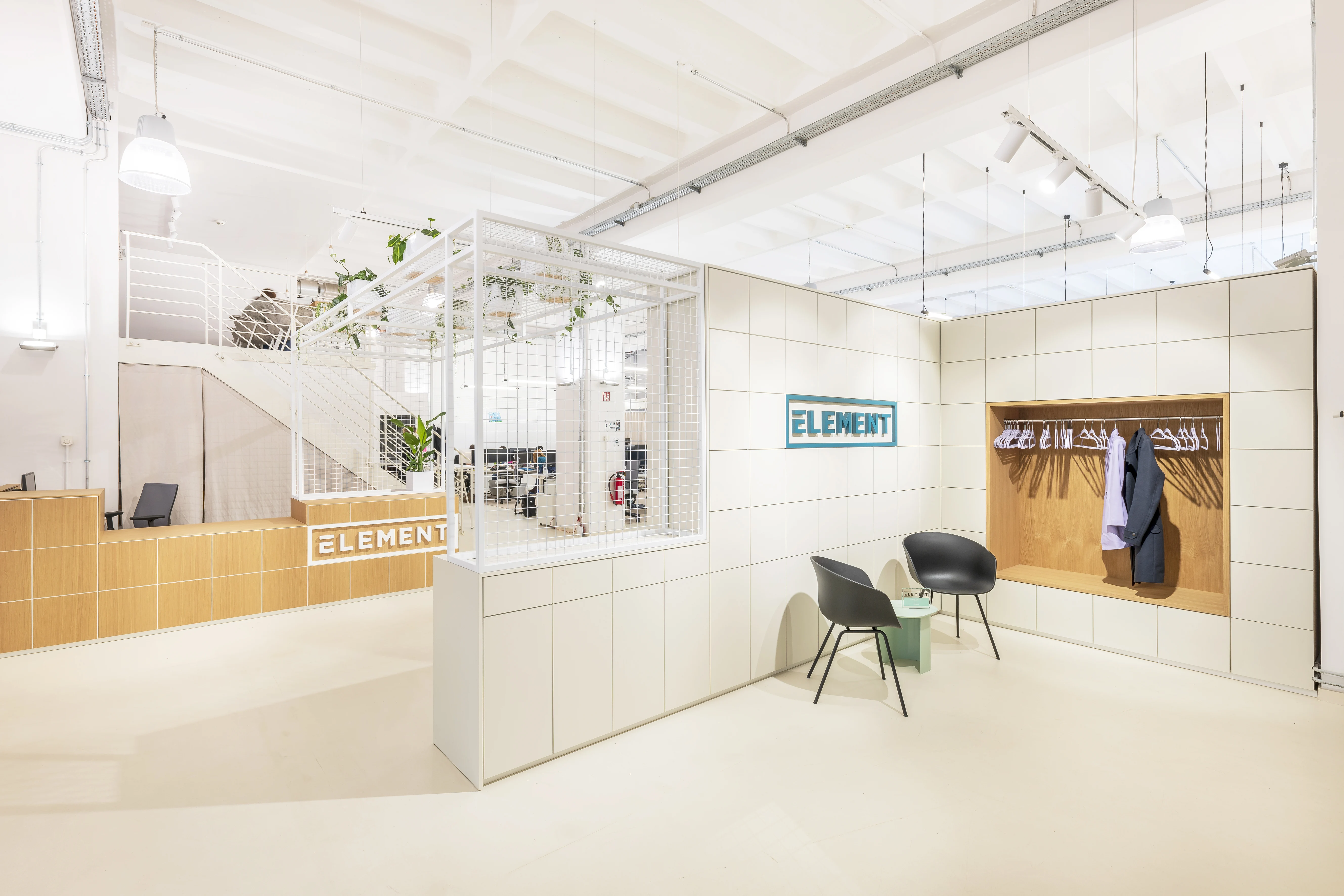 Entrance zone at ELEMENT with a sand-coloured tiled reception counter, white grid partitions with hanging plants, the ELEMENT logo on a tiled wall and an oak coat niche with two black chairs.