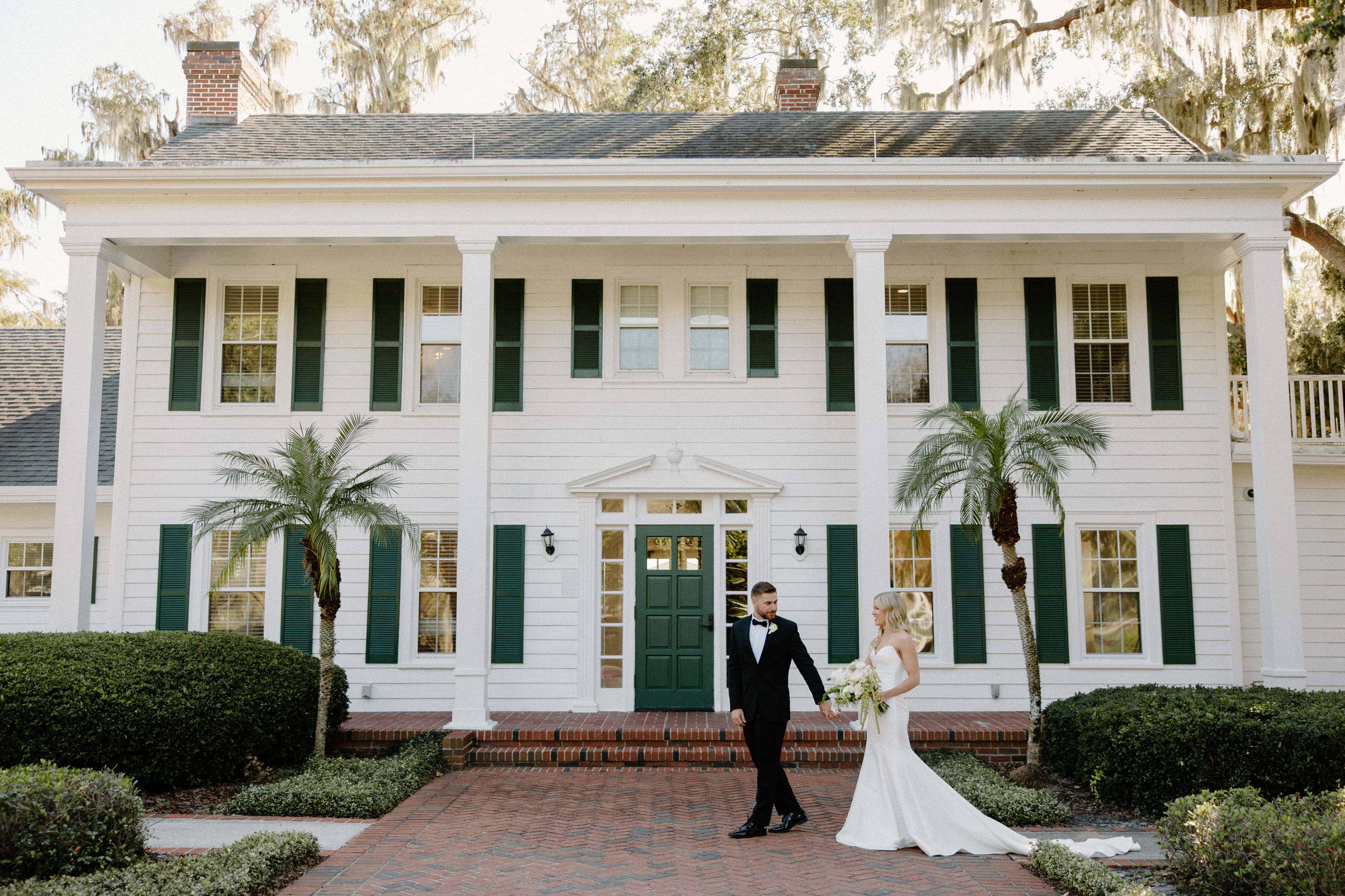 A wedding couple walks hand-in-hand in front of a large, white plantation-style house with green shutters.