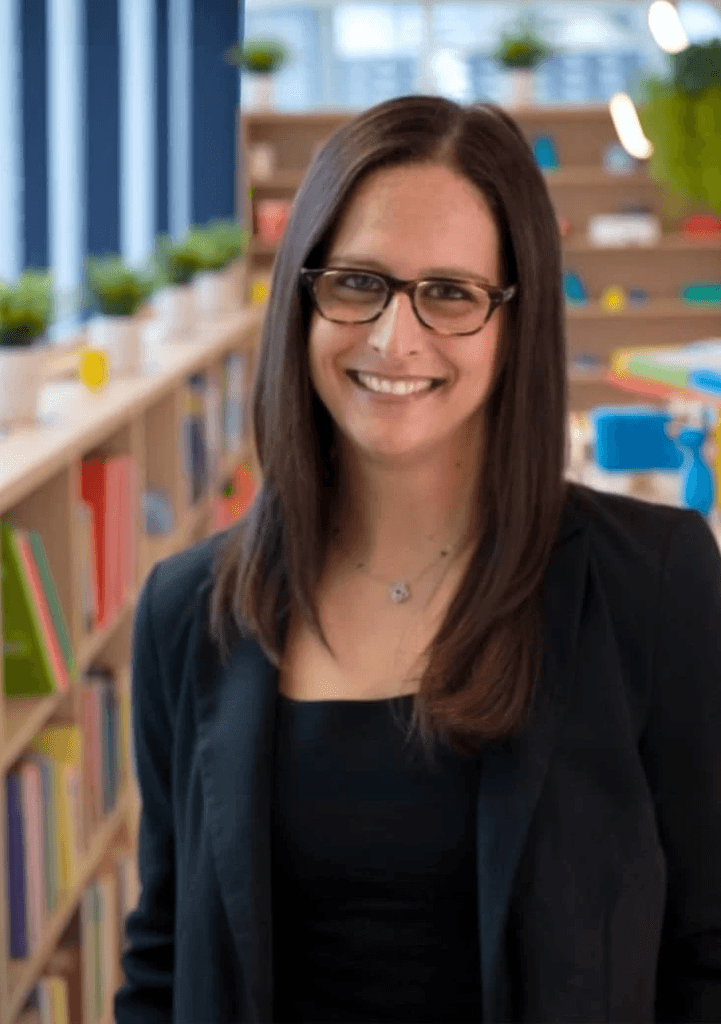Risa Battino, owner and speech-language pathologist at Communication Connect, smiling in a colorful, book-filled therapy space.
