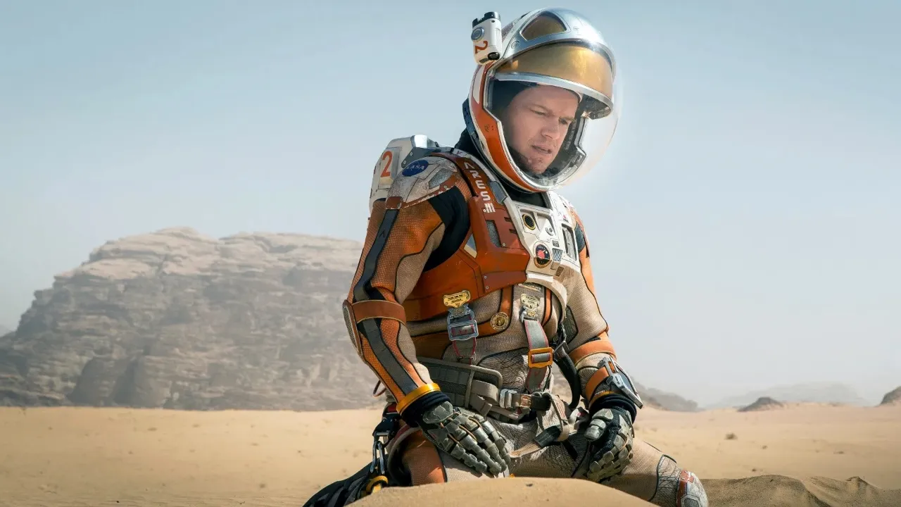 A man in a high-tech astronaut suit is kneeling on a sandy, rocky terrain resembling the Martian landscape, under a clear blue sky with distant mountains visible in the background.