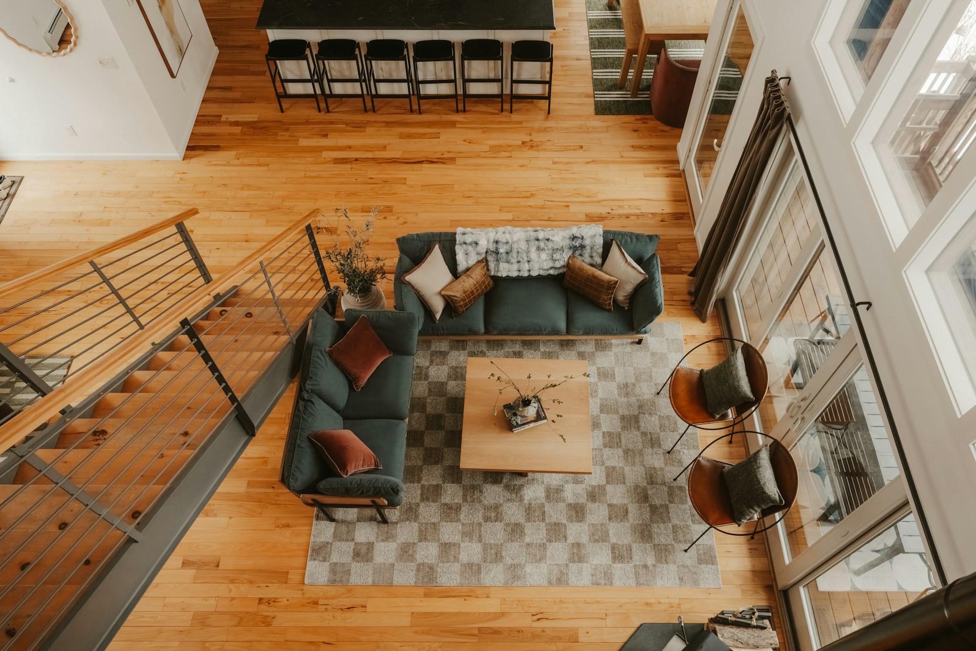 High-angle view of a cozy living room with green sofas, wood floors, a checkered rug, stairs, and a kitchen bar.