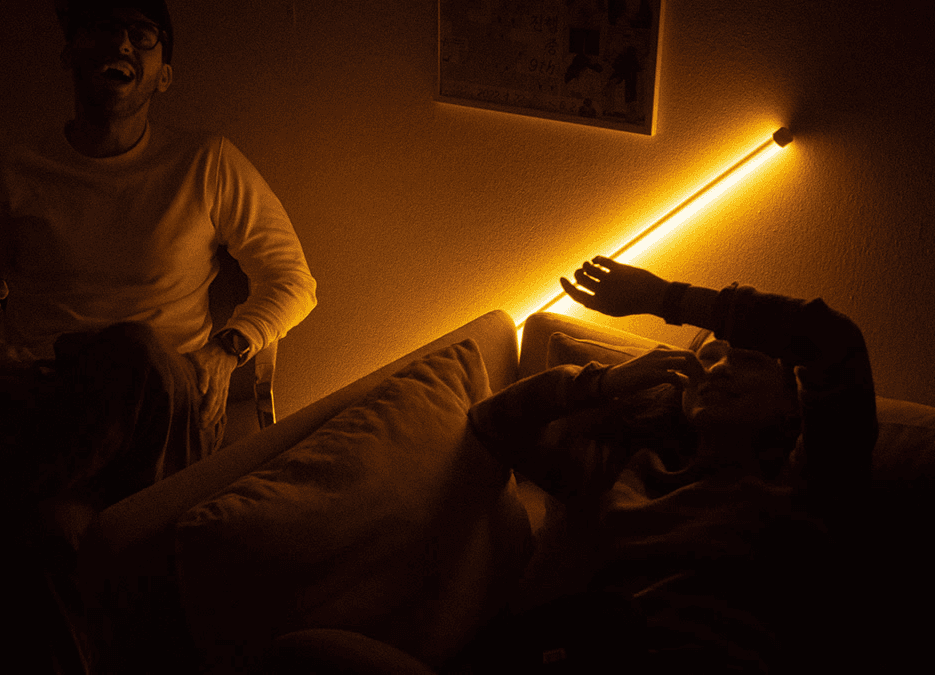 An intimate, low-light shot of guests relaxing on a sofa during the studio opening party, illuminated by a warm, linear amber light fixture on the wall. This image captures the cozy, inviting atmosphere of the new office space and the studio’s focus on human-centric design and community.