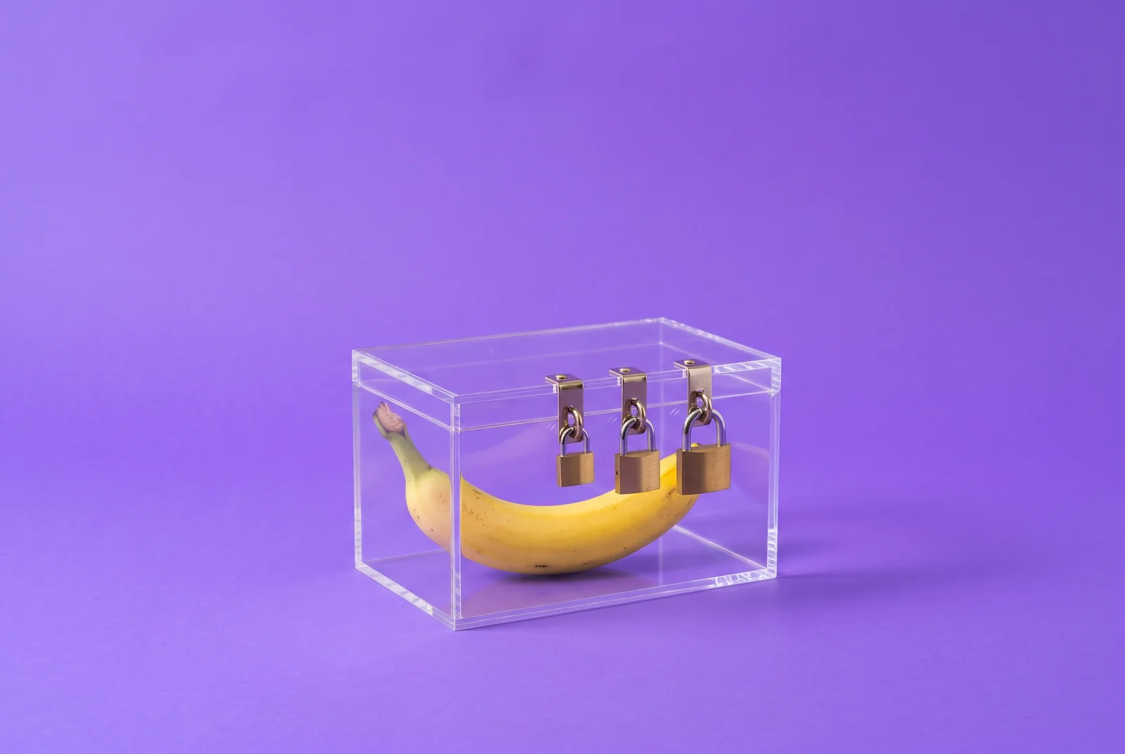 A banana locked inside a clear acrylic box with three padlocks of different sizes, a playful image for how to block adult sites on your phone permanently