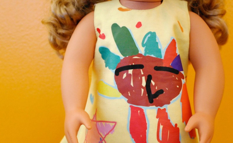 A doll with curly brown hair wears a pale yellow sleeveless top decorated with colorful paint splatters. The doll’s face is drawn with black lines for eyes, eyebrows, and a mouth, giving it a whimsical appearance. The background is a solid orange-yellow color.