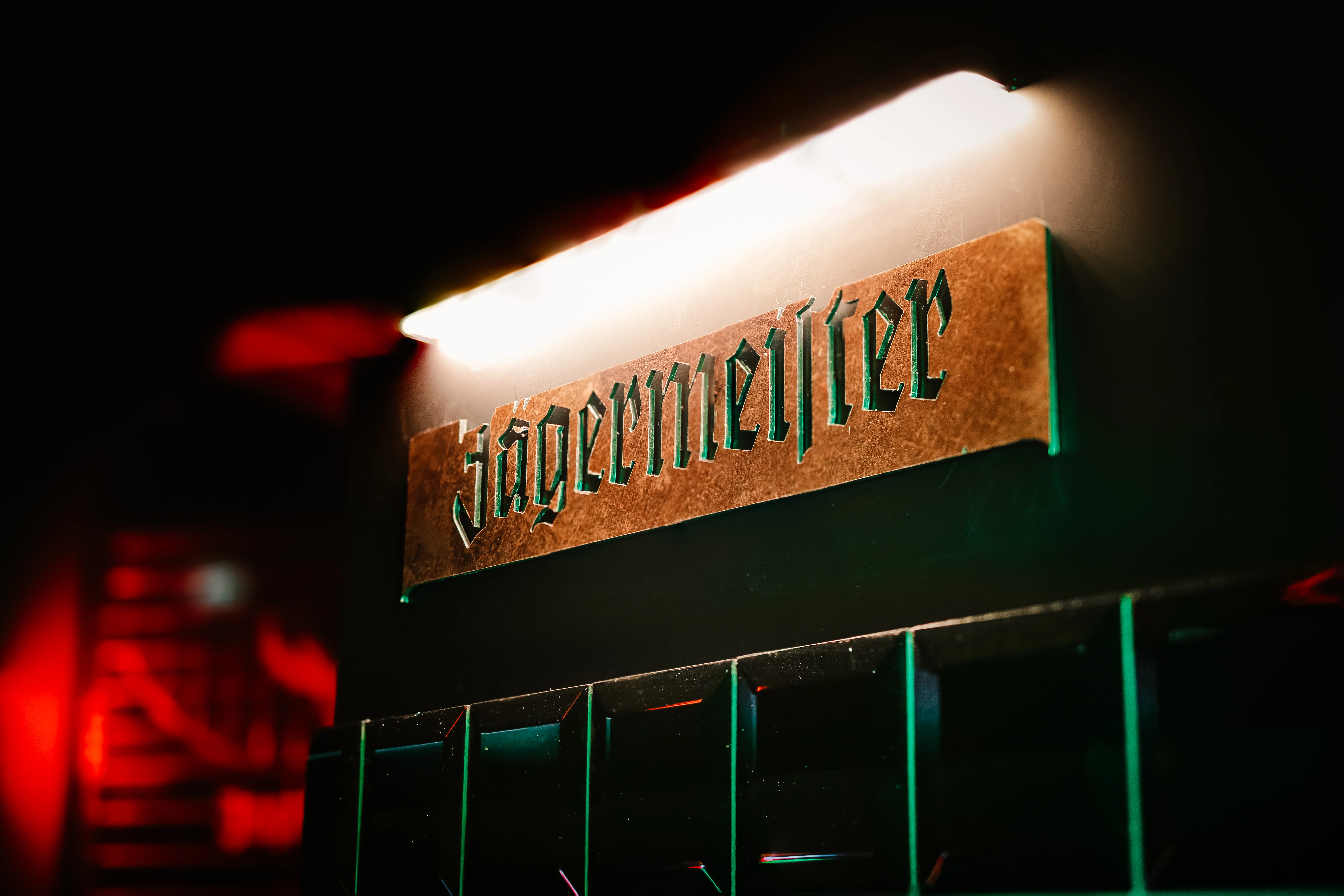 Copper Jägermeister sign in gothic lettering mounted on a dark wall at JÄGERMEISTER x CALL ME DRELLA, lit by a warm tube light from above.