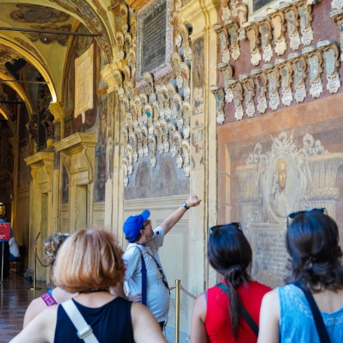 A tour group looks at intricate wall carvings and murals inside an ornate historical building. The guide points at details.