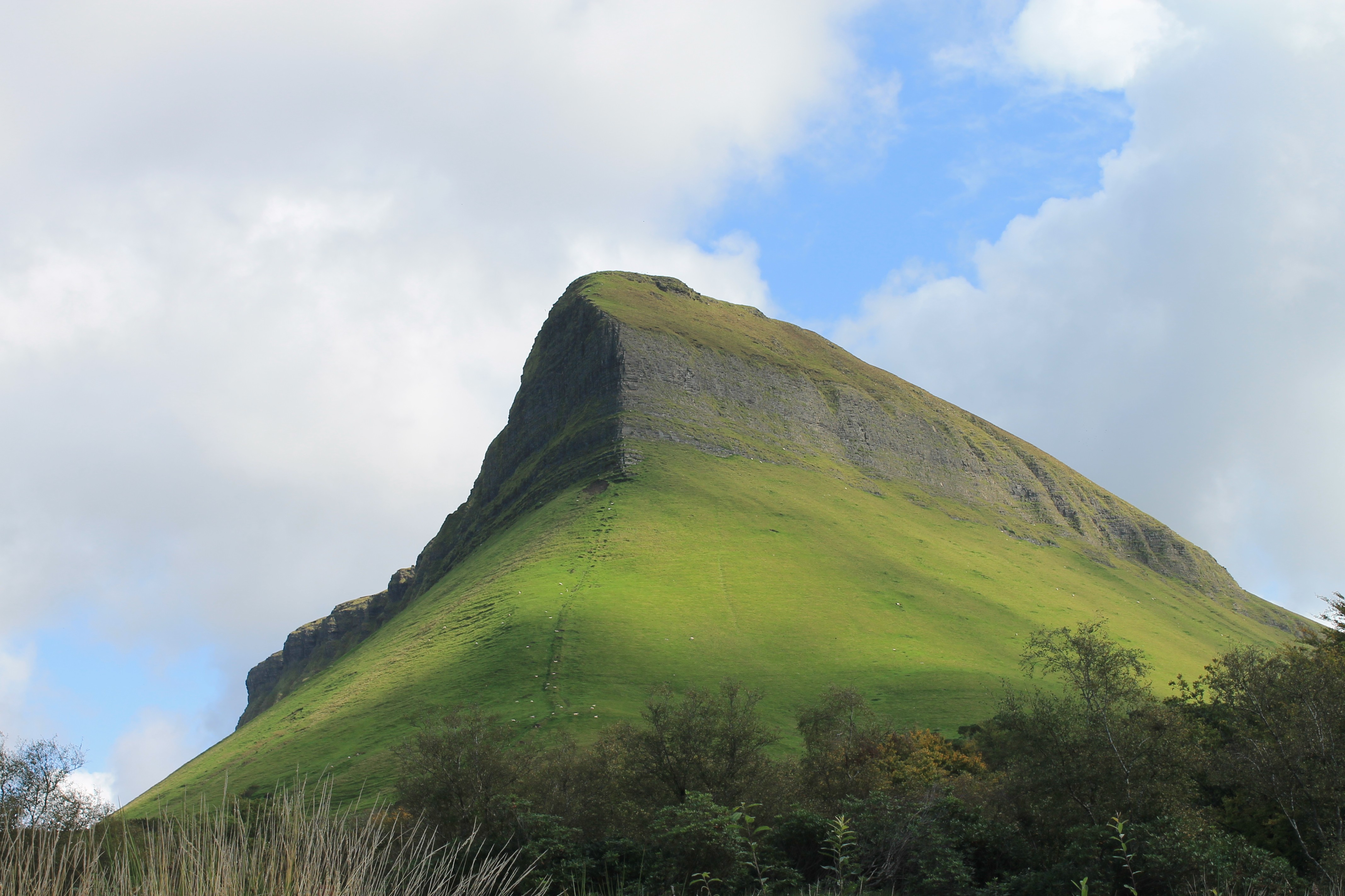 A green, steep mountain peak rises against a blue sky with white clouds.