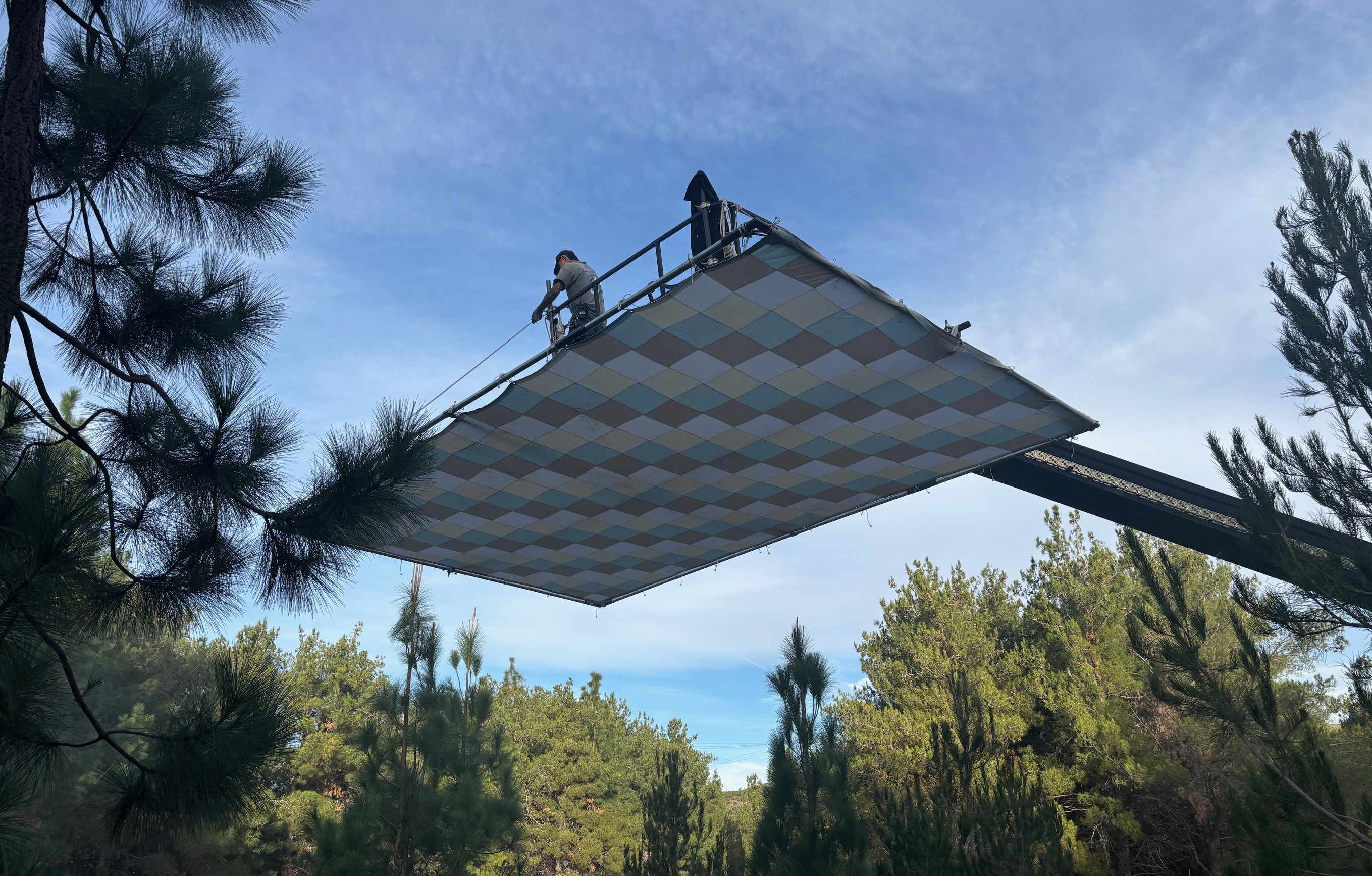 12x20 checker bounce flat overhead with an operator visible on top of the frame, daylight underneath, pine trees at the lower edge.