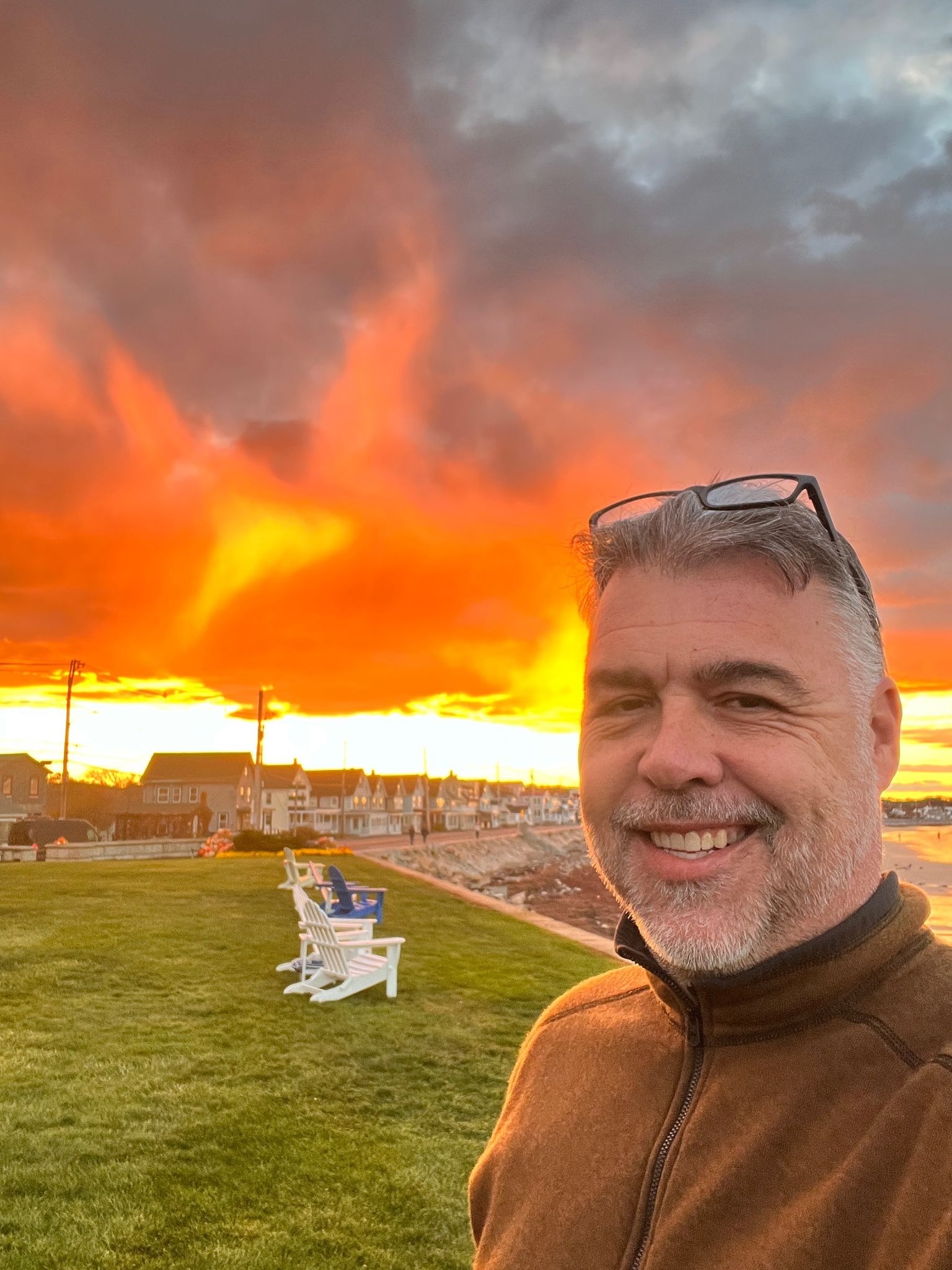 Man smiling at fiery beach sunset with colorful clouds and seaside homes in background.