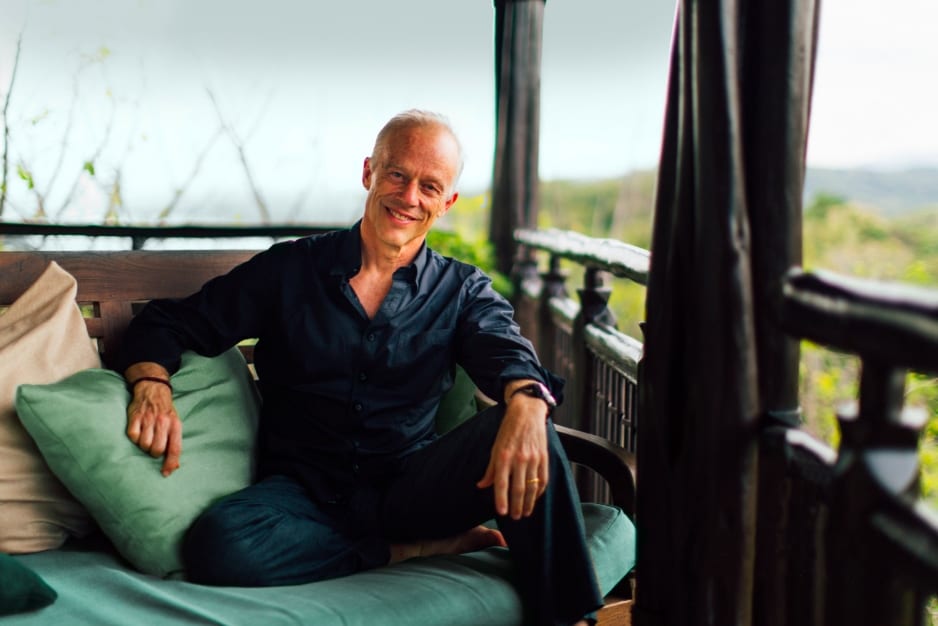 Man in black shirt relaxing on outdoor balcony bench with pillows, lush greenery in background.