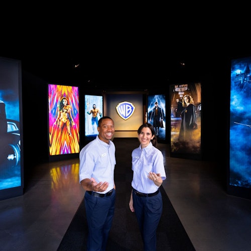 Two people in uniform stand in an exhibit hall with large posters of movie characters and a Warner Bros. logo in the background.