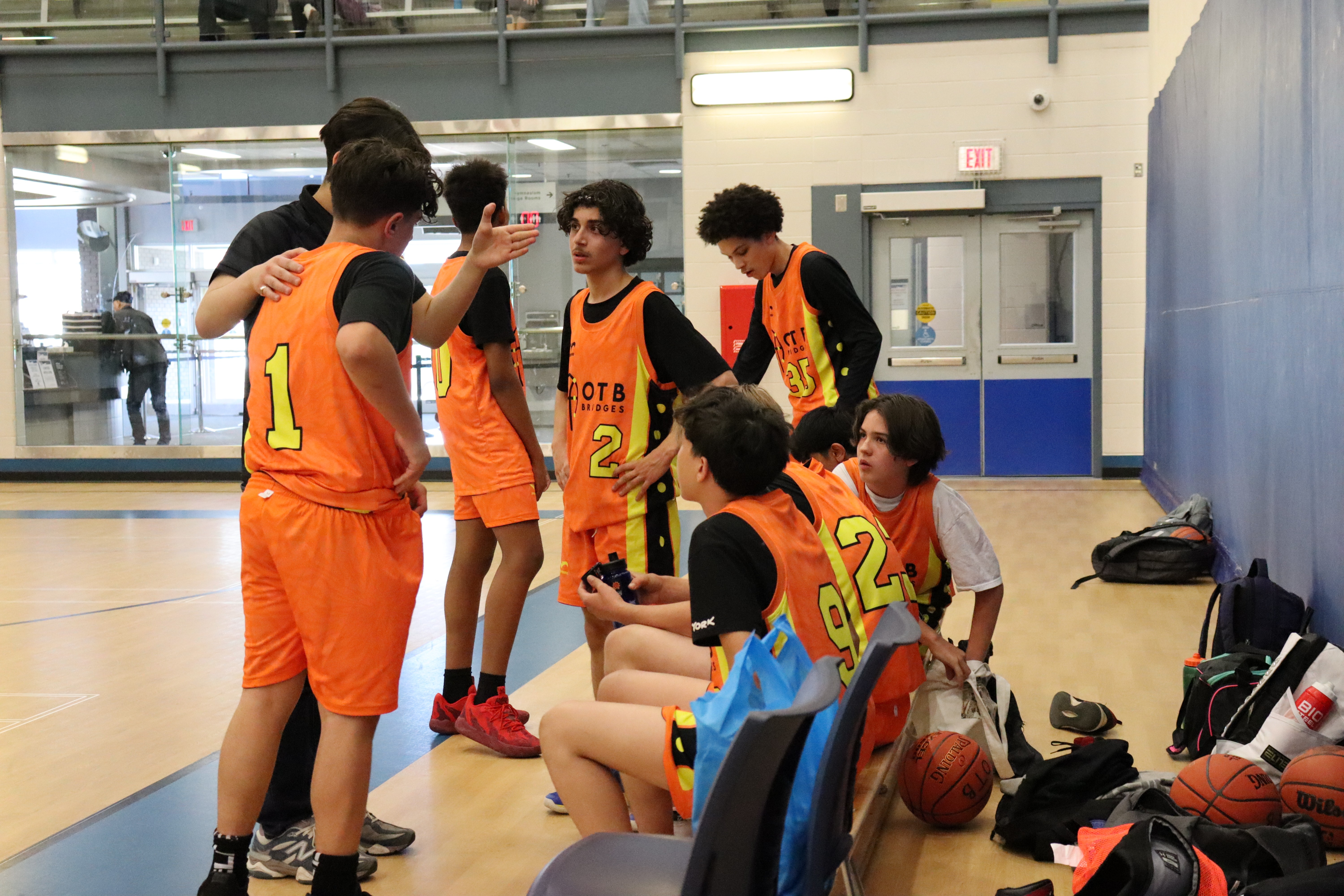 A youth basketball player in an orange jersey dribbling aggressively past a defender during a game, with a referee and spectators visible in the background on an indoor court.