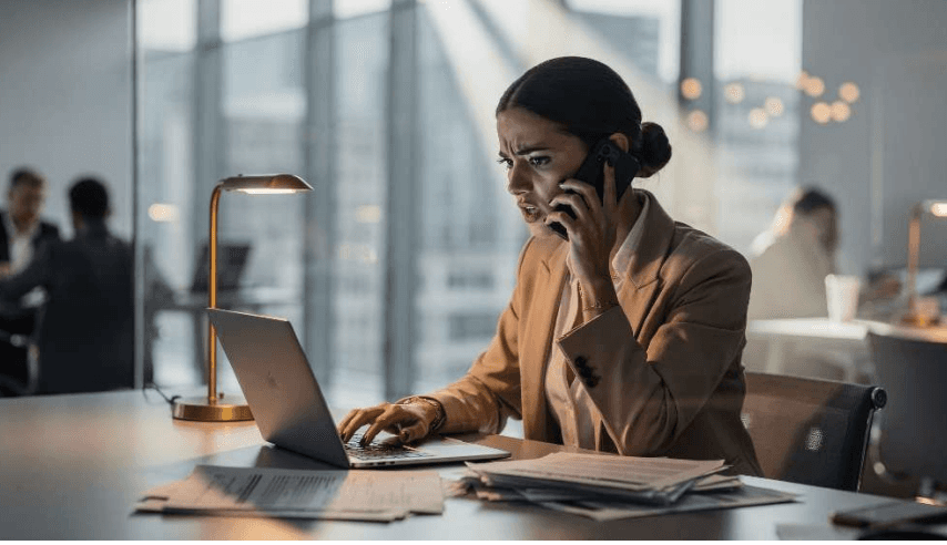 Frau im Business-Outfit sitzt abends im Büro am Laptop, telefoniert konzentriert und wirkt angespannt, während Dokumente auf dem Schreibtisch liegen.