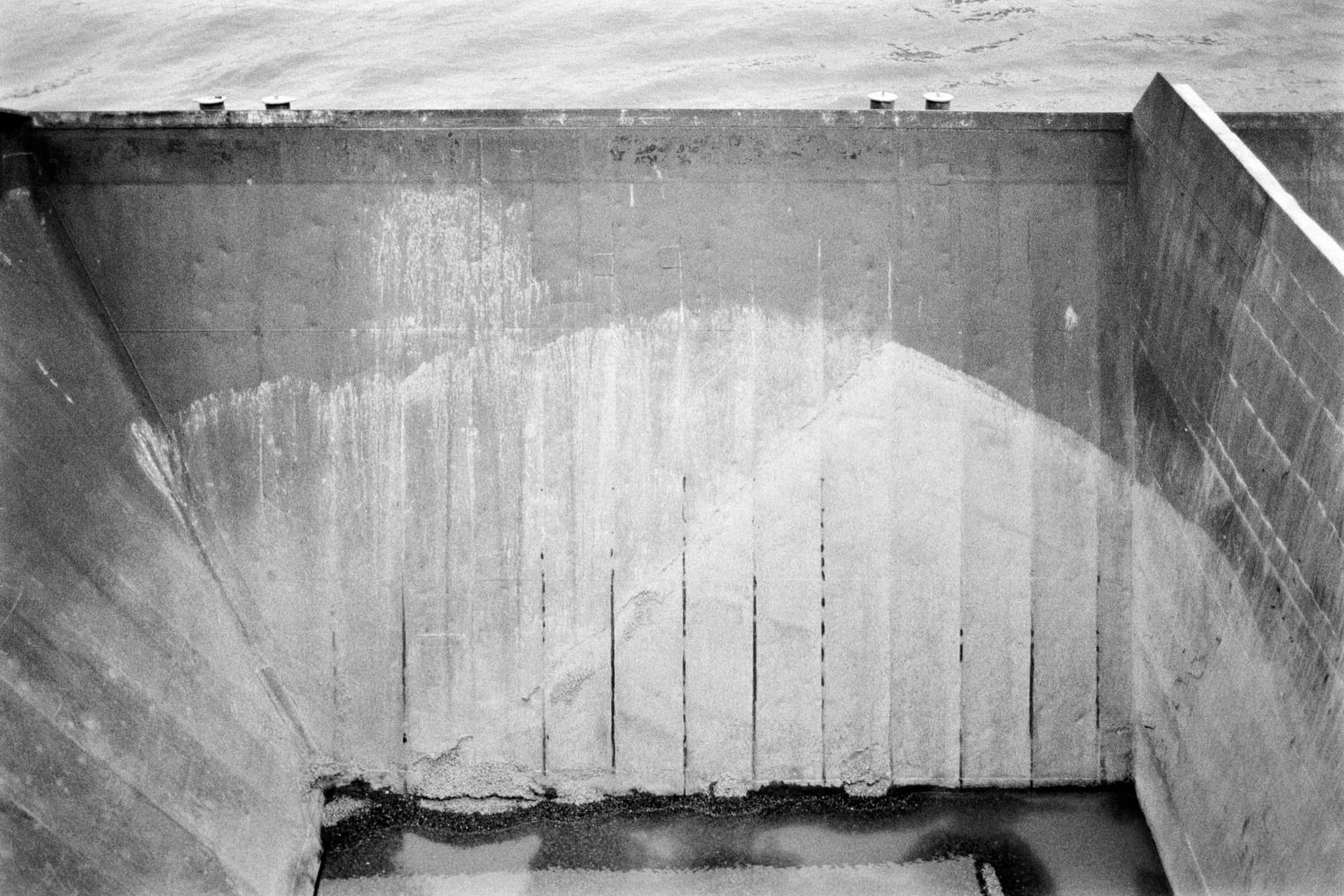 Empty ship or vessel interior hold viewed from above, weathered steel walls with water staining and vertical weld lines, sand and debris accumulated at base