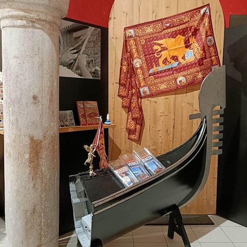 Una replica di una gondola esposta al coperto, con libri a tema veneziano e un arazzo su una parete di legno sullo sfondo.