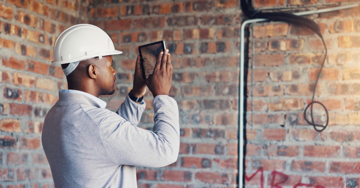 A man wearing a white hard hat points a tablet toward a brown brick wall with some graffiti on it. He takes a photo of the wall.