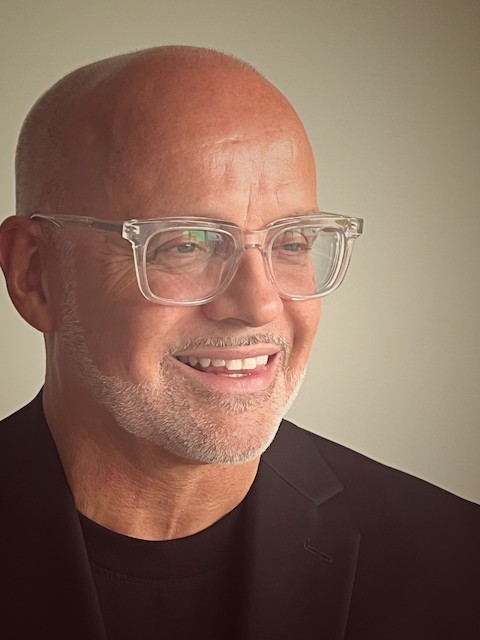 Portrait of Stéphane Paquet, smiling, wearing clear glasses, a black blazer, and a black shirt.