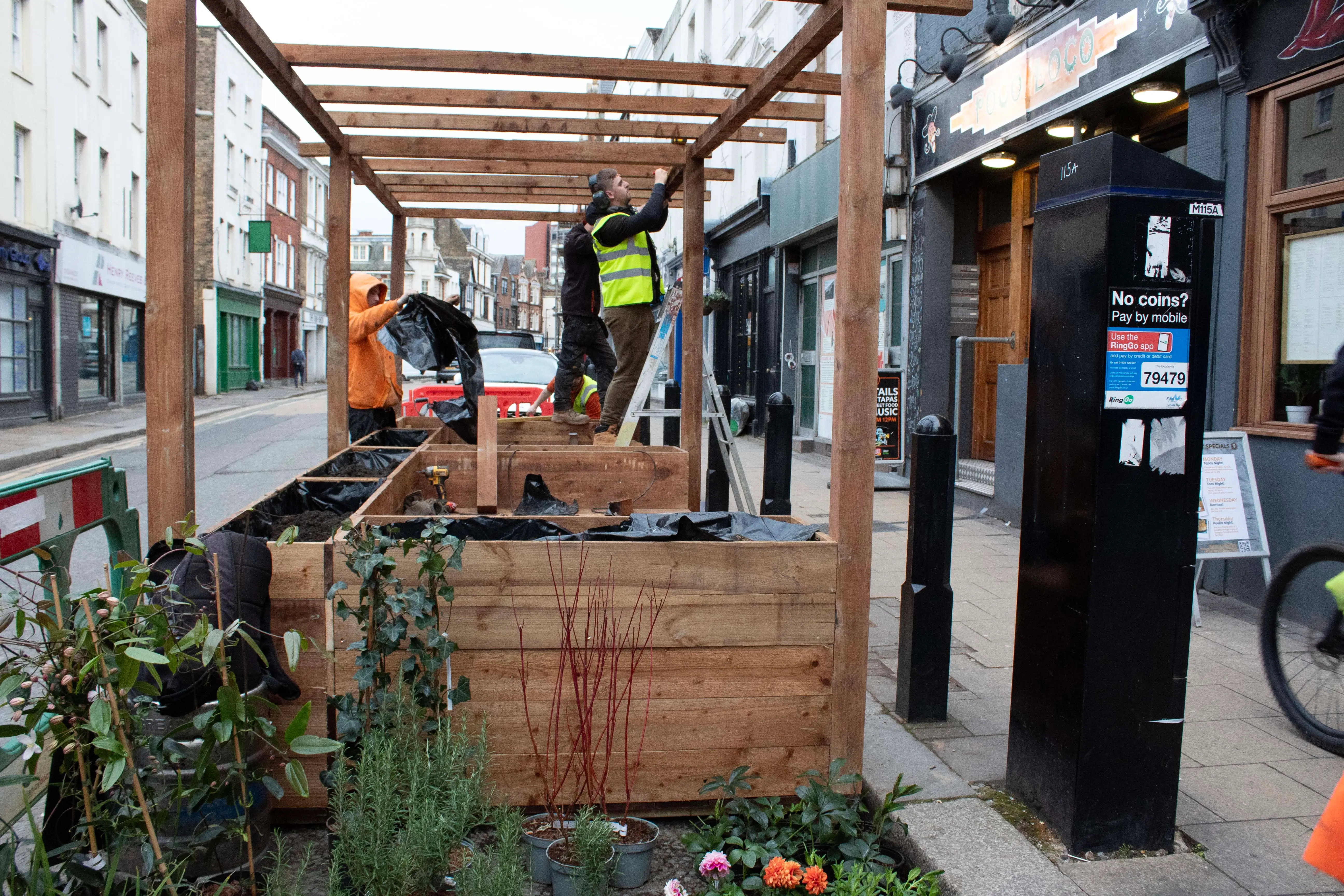 A wooden structure with plants, outdoor seating, and people working, in an urban setting.