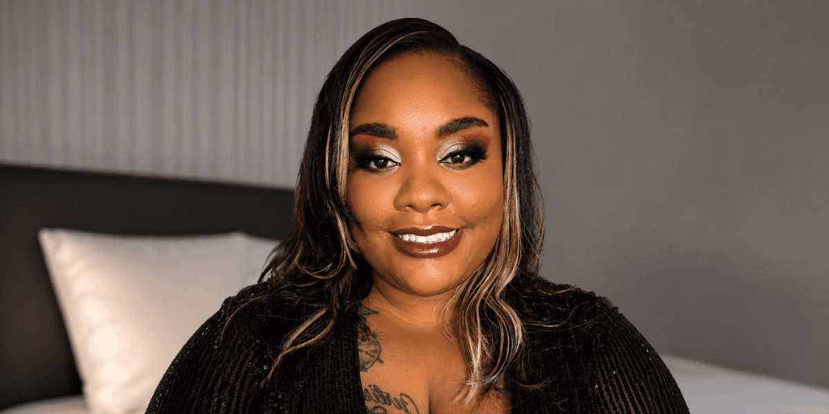 Woman with styled hair and bold makeup smiles in black sequined outfit, seated in softly lit bedroom.