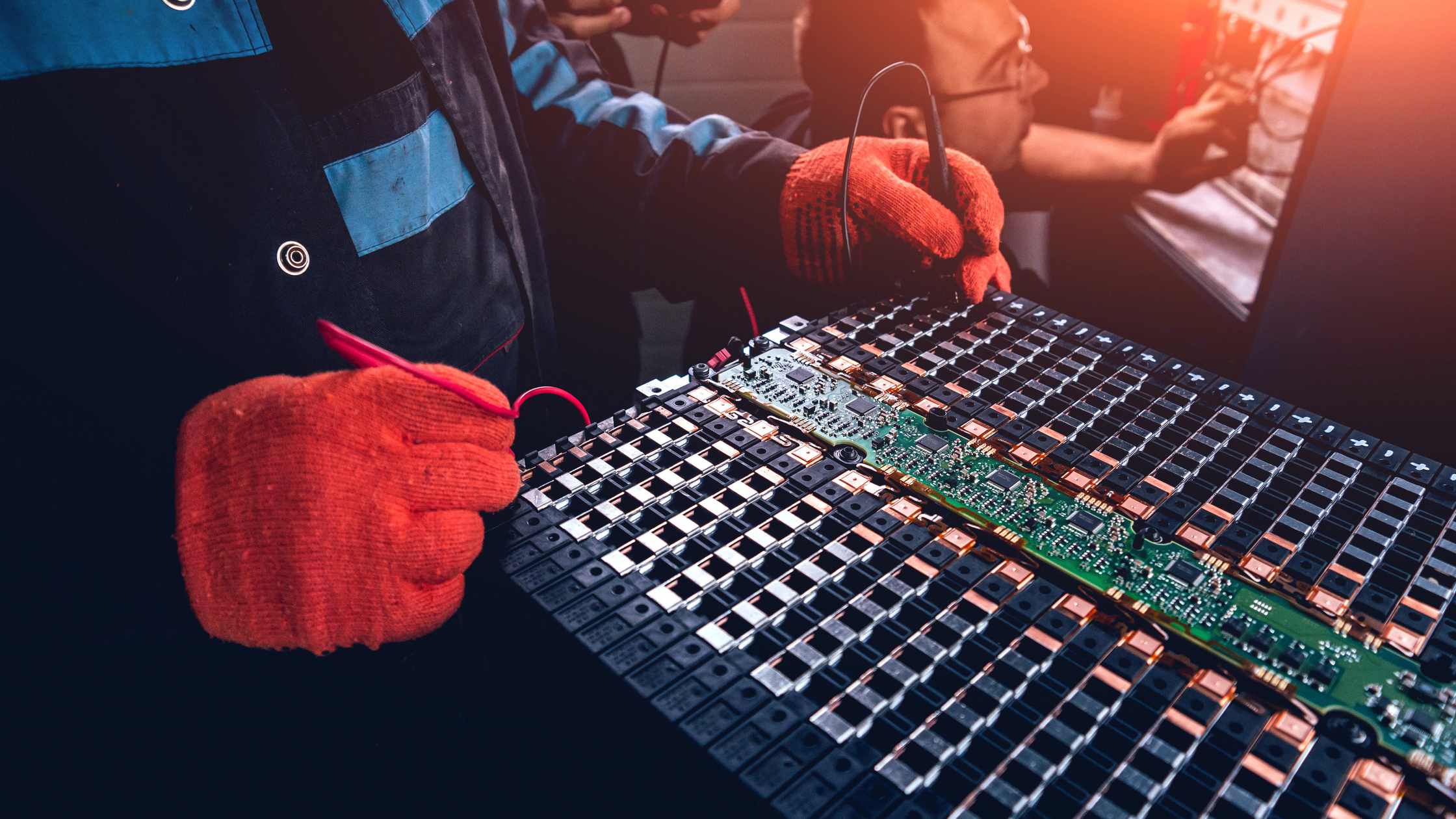 Workers in orange gloves using multimeter probes to test a large electric vehicle battery module