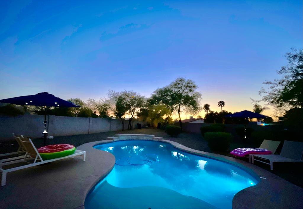 A tranquil evening view of an affordable luxury vacation rental house's swimming pool, surrounded by lounge chairs and illuminated by the soft glow of outdoor lights under a twilight sky.