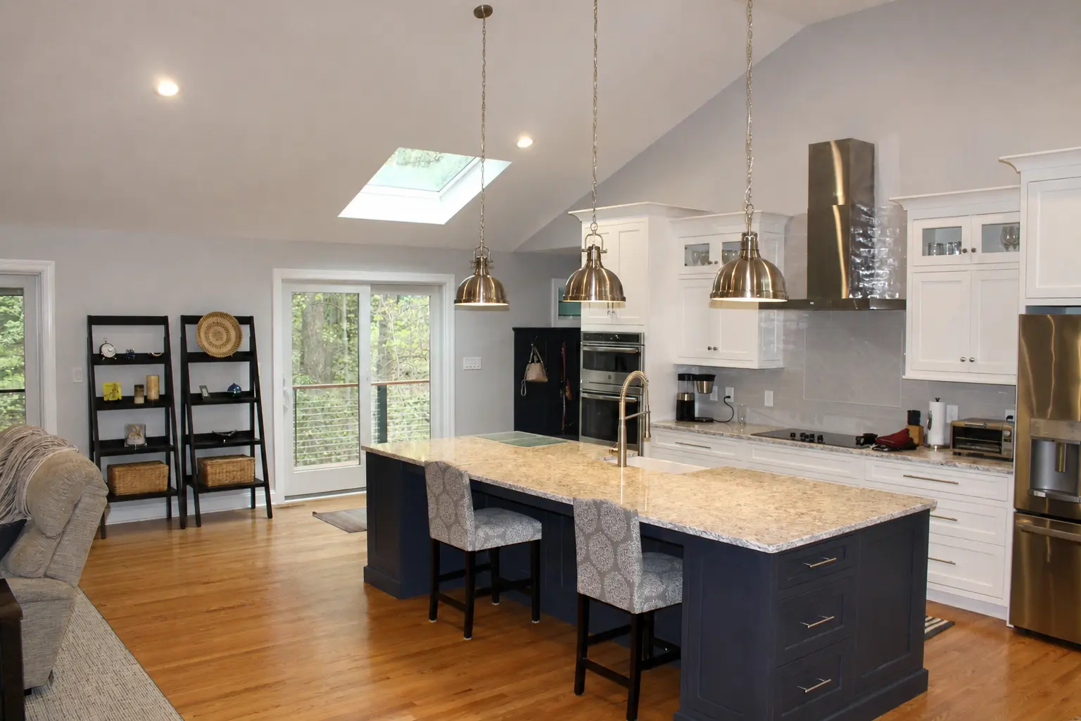 Modern renovated kitchen with white cabinetry, dark navy island, granite countertops, stainless steel appliances, pendant lighting, hardwood floors, and vaulted ceiling with skylight.