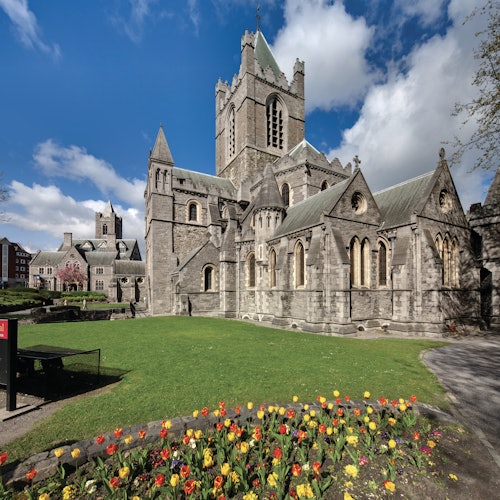 Stone cathedral with multiple towers surrounded by green lawn and colorful flowers, under a bright blue sky with scattered clouds.